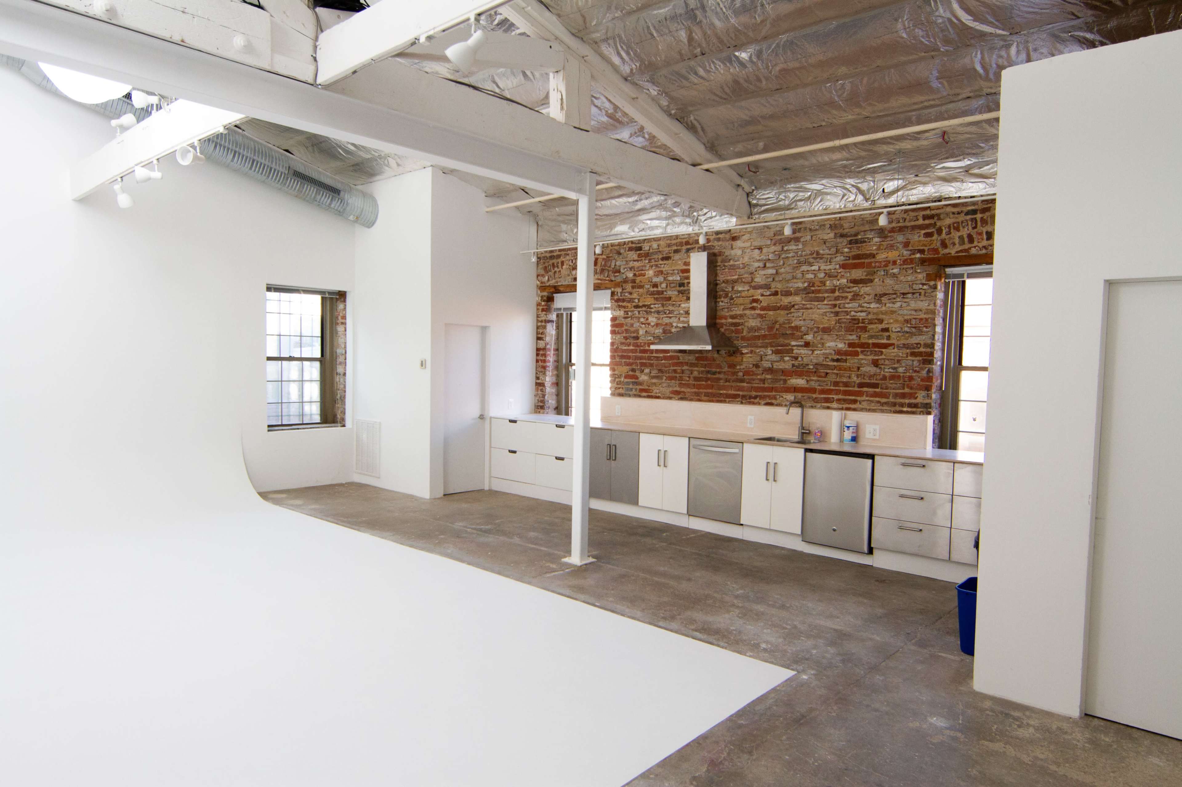 A spacious studio with a large white backdrop on one side and a kitchen area with brick walls and stainless steel appliances on the other.