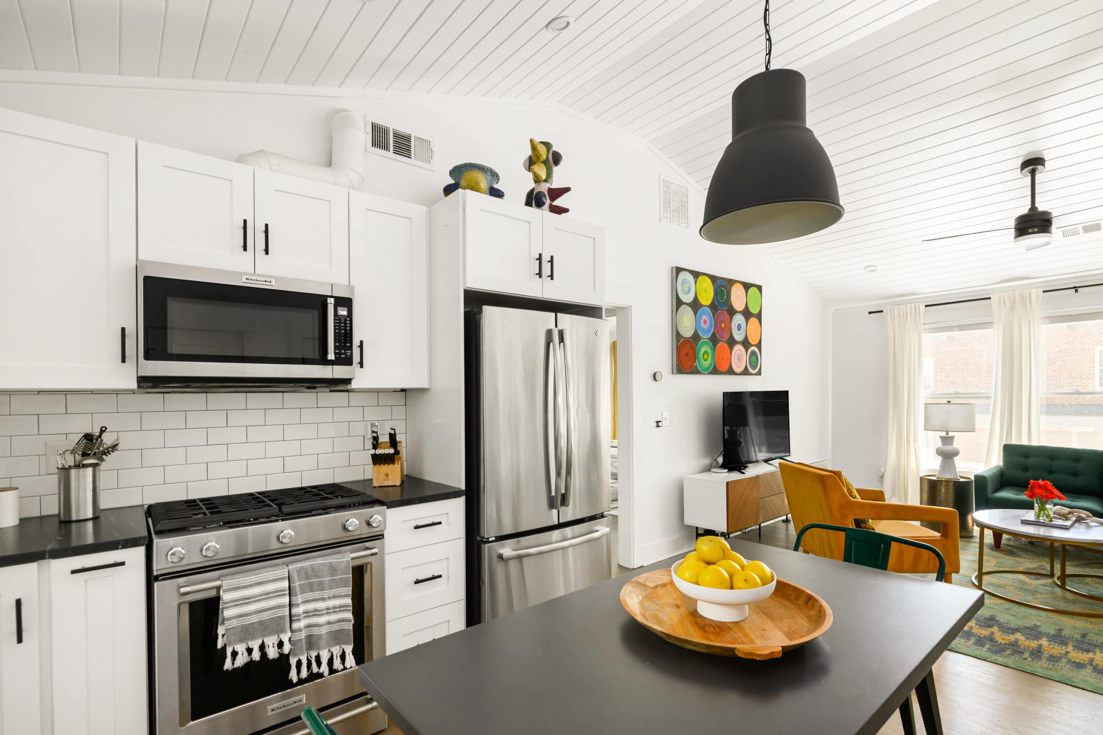 The image shows a modern kitchen featuring white cabinets, stainless steel appliances, and a black countertop, with a dining area that includes a round table and a bowl of lemons.