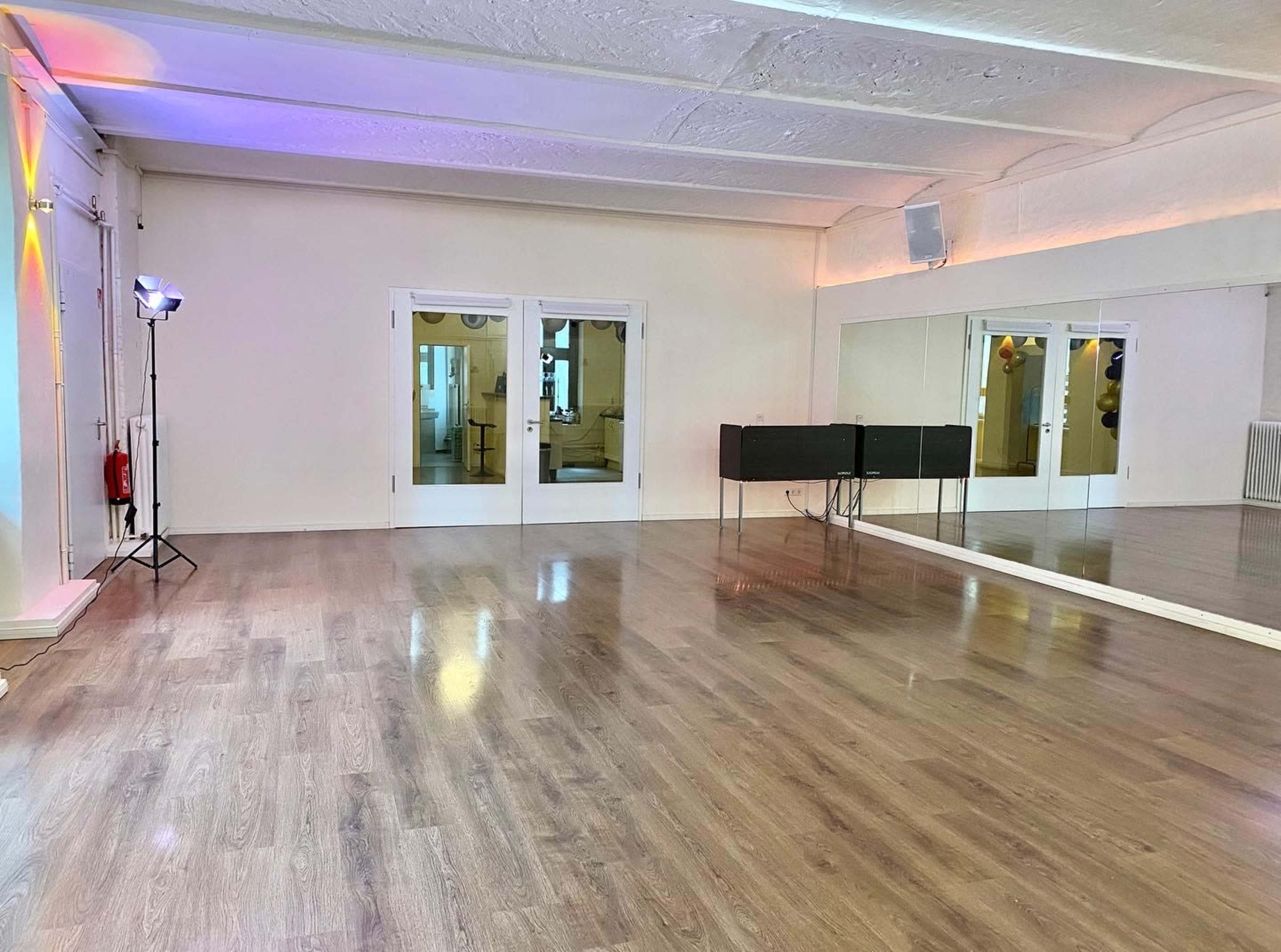 An empty dance studio with wooden flooring, a large mirror on one wall, and glass doors leading to another room.