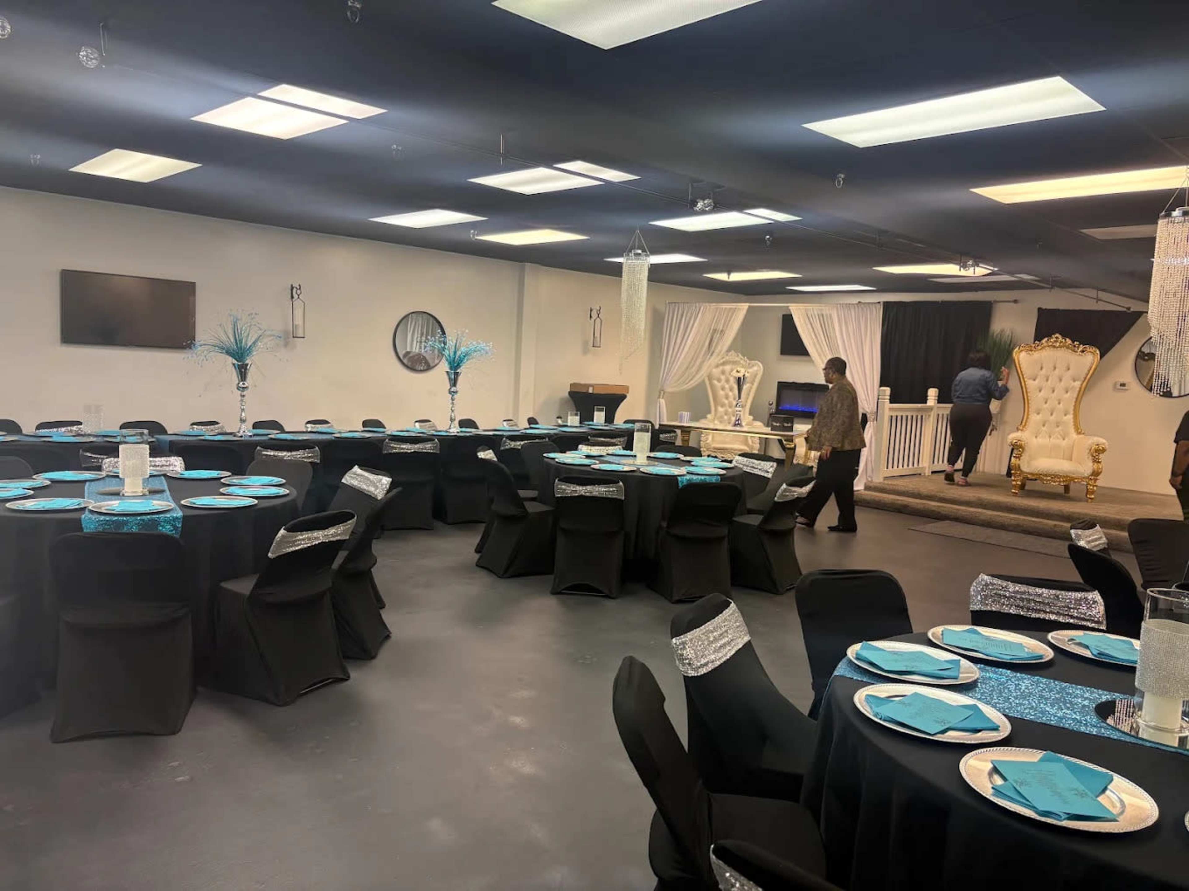 The image shows a decorated event space with tables set for a formal gathering, featuring black tablecloths and turquoise accents, alongside elegant seating at the back.