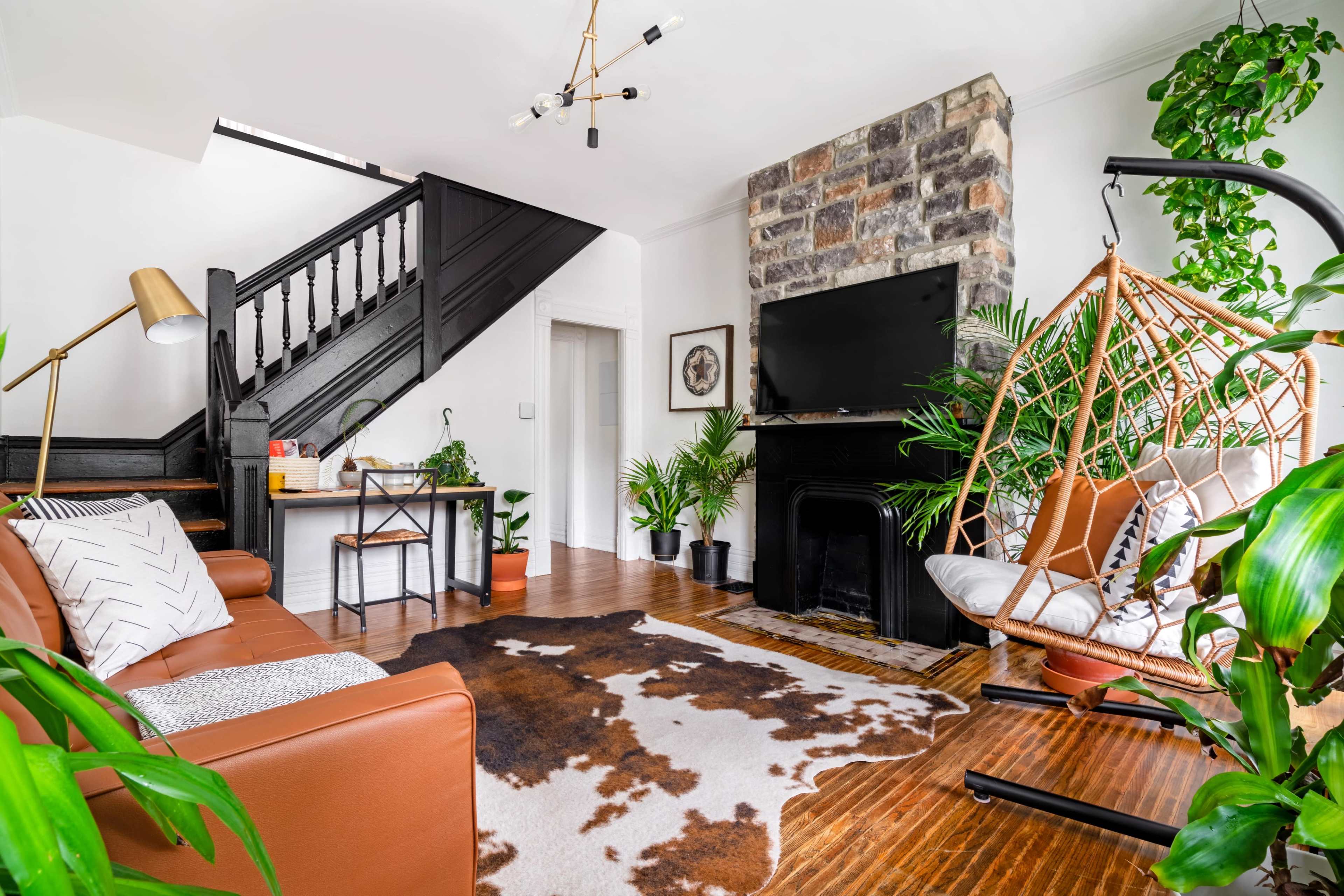 A modern living room features a brown leather sofa, a large cowhide rug, a black fireplace, and an assortment of indoor plants, complemented by a staircase and a television mounted above the fireplace.
