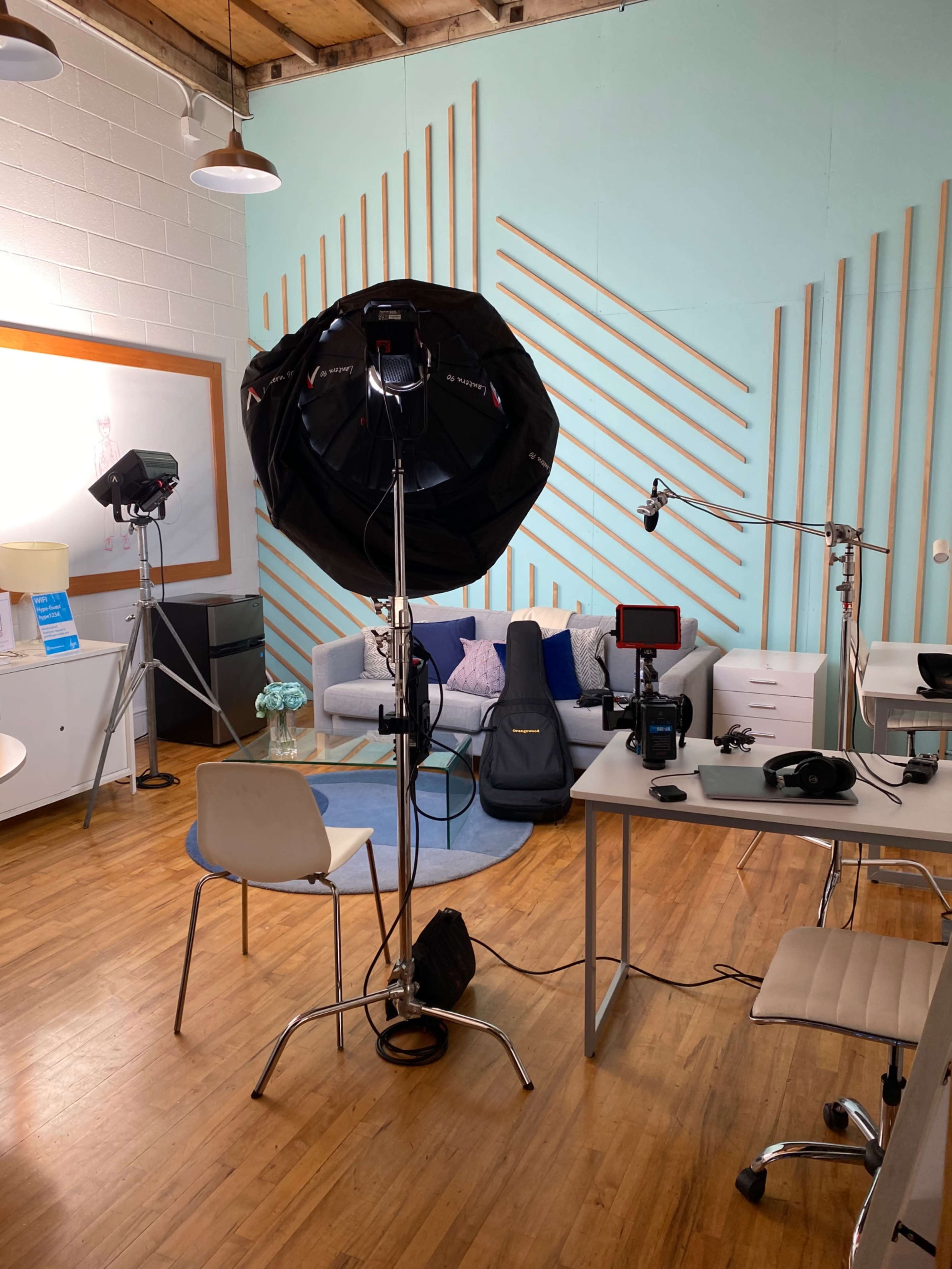 The image shows a well-lit indoor studio setup featuring a sofa, a desk with equipment, and professional lighting gear.