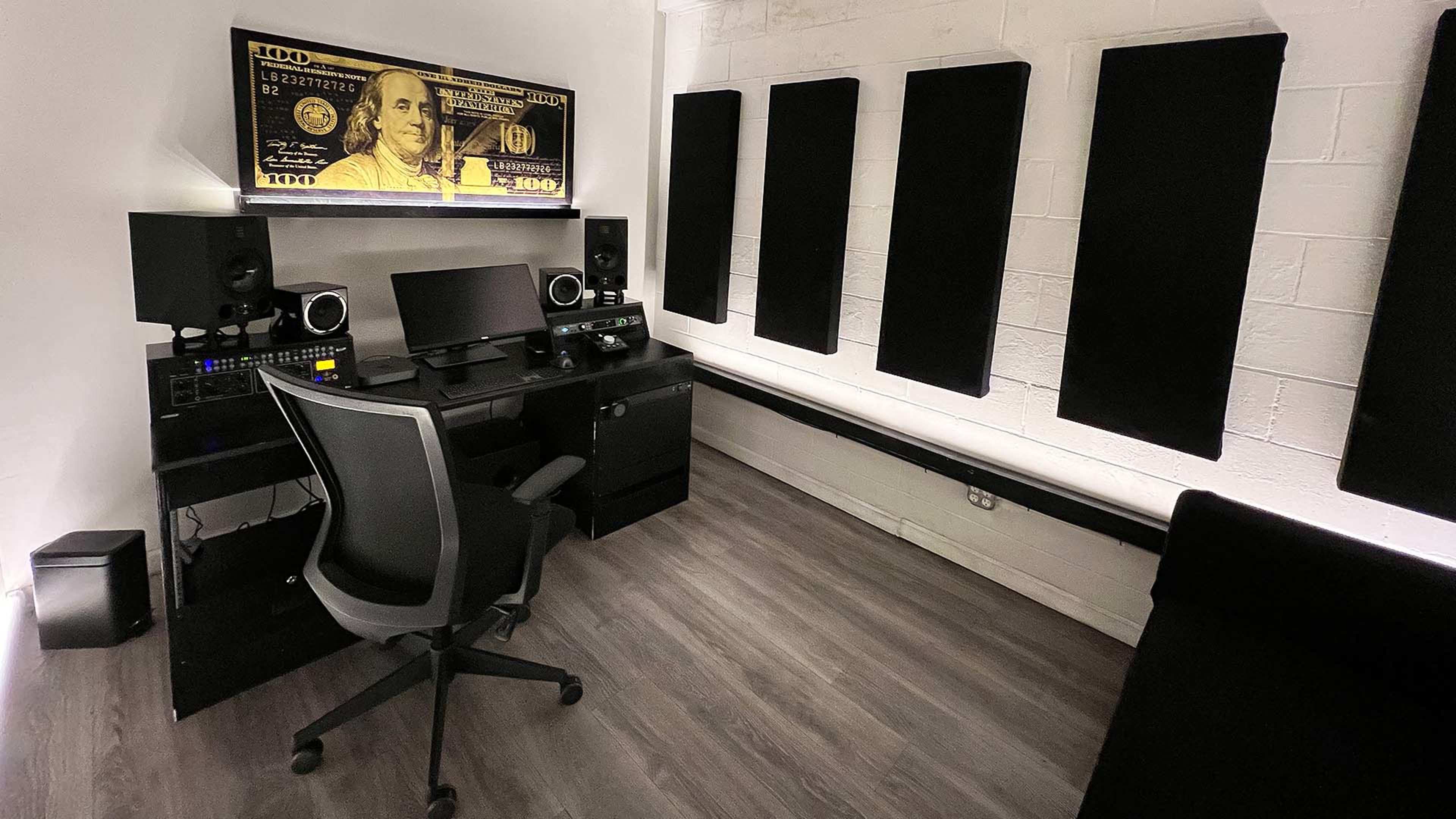 A modern workspace with a desk, computer setup, and acoustic foam panels on the walls, featuring a large depiction of a hundred-dollar bill above the monitor.