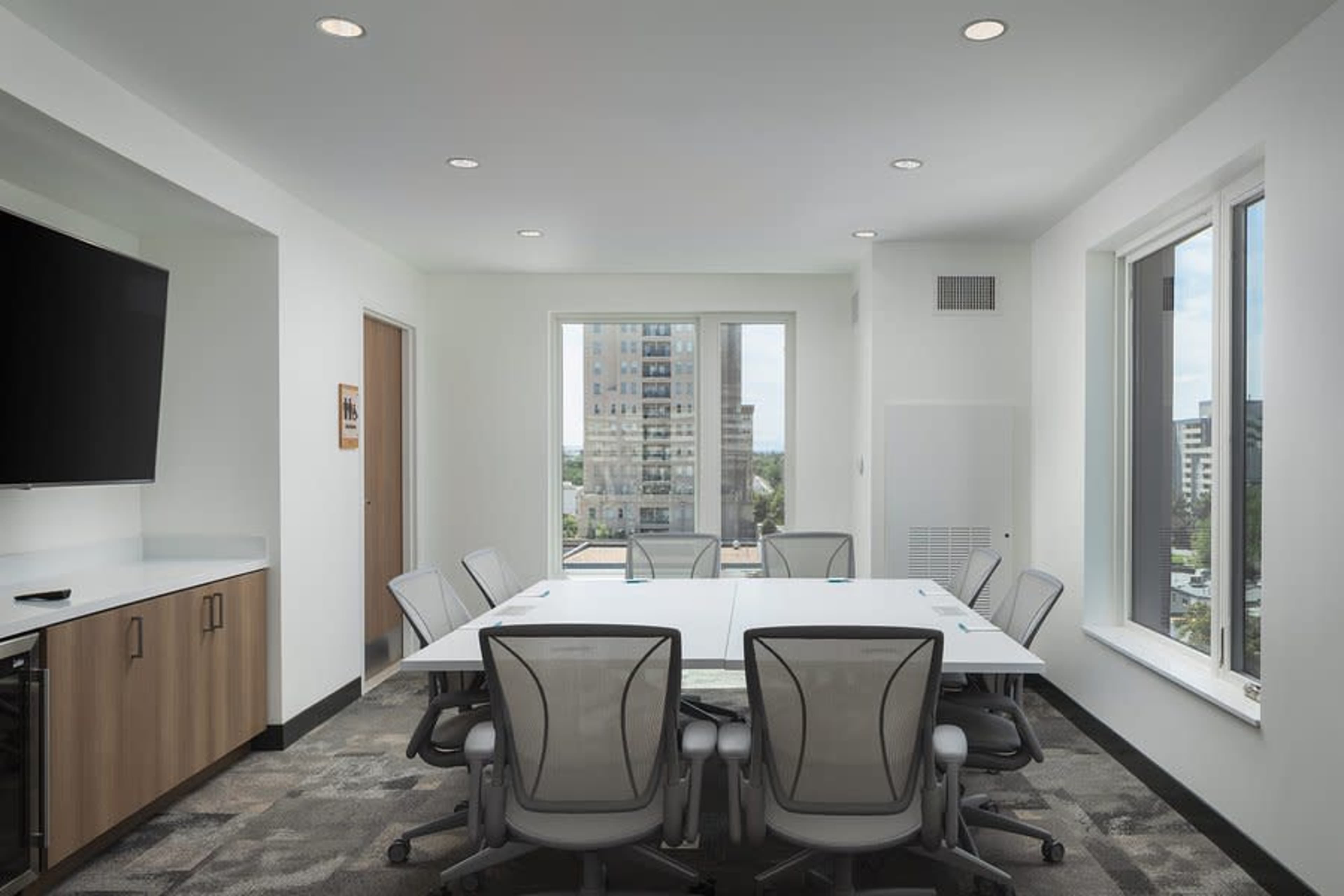 A modern conference room features a large white table surrounded by ergonomic chairs, with windows providing a view of a cityscape.
