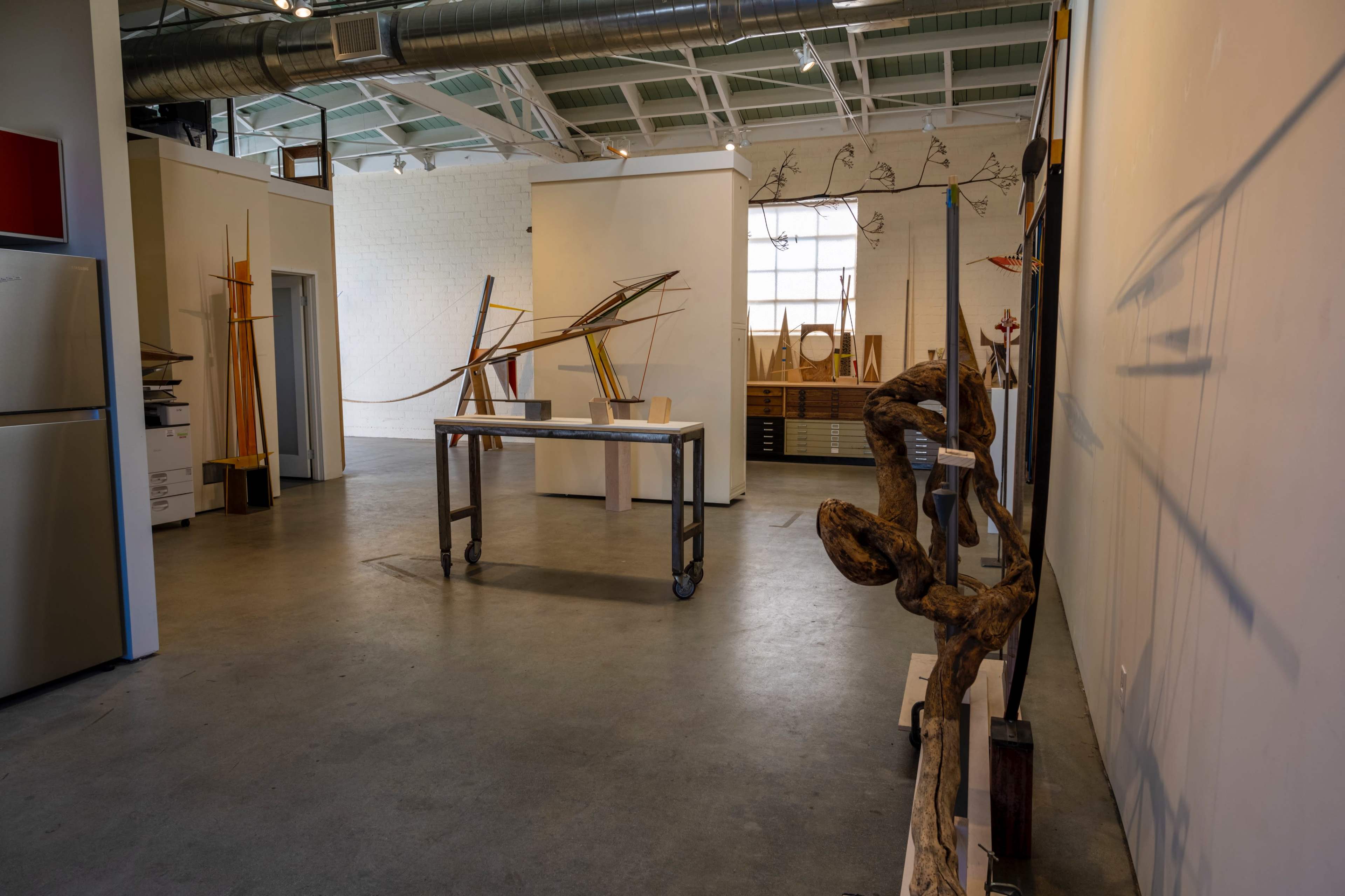 An art gallery features various sculptures displayed on pedestals and stands, with large windows providing natural light.