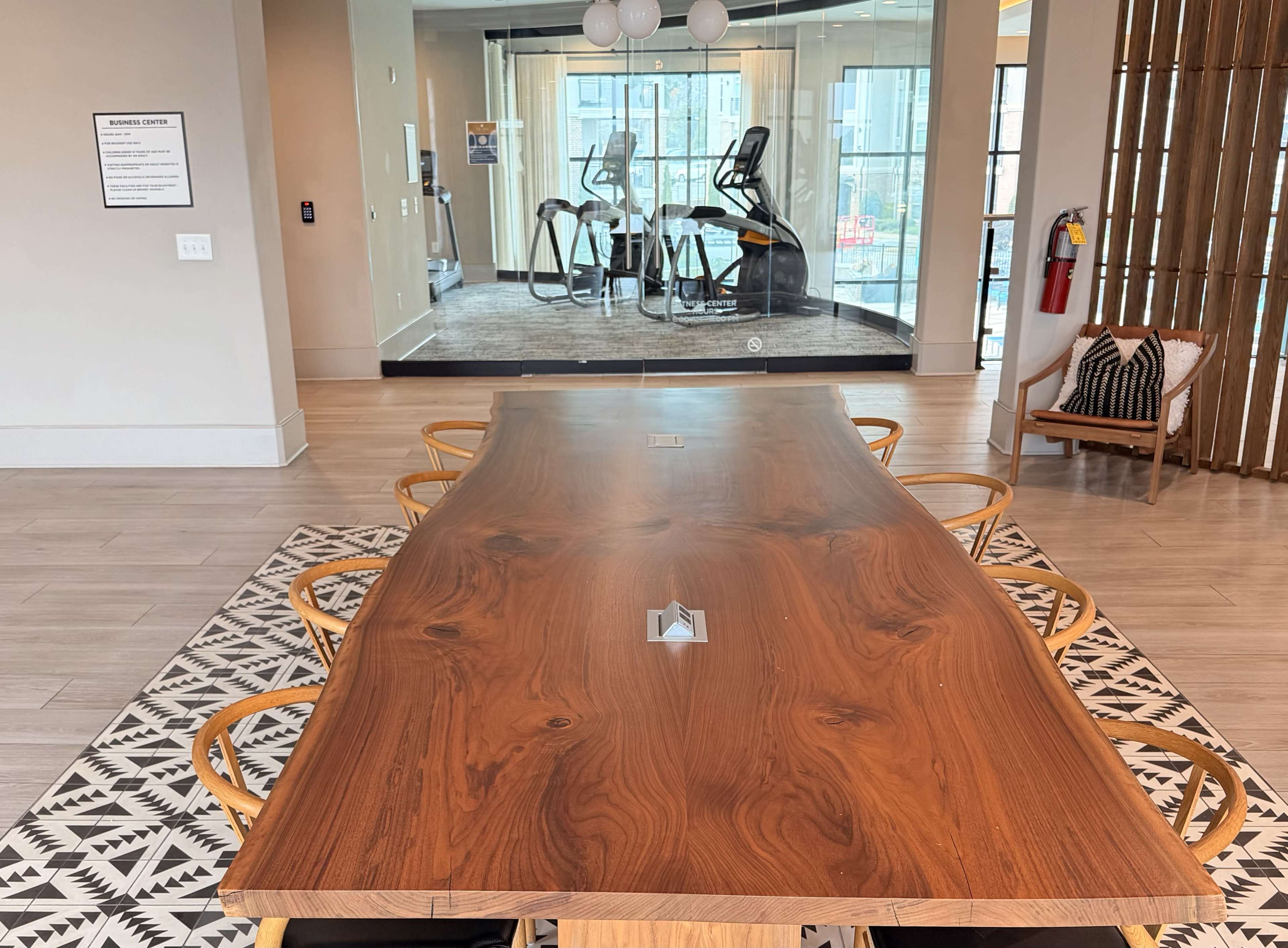 A large wooden table with several chairs surrounds it, located in a room adjacent to a gym area featuring exercise equipment visible through glass walls.