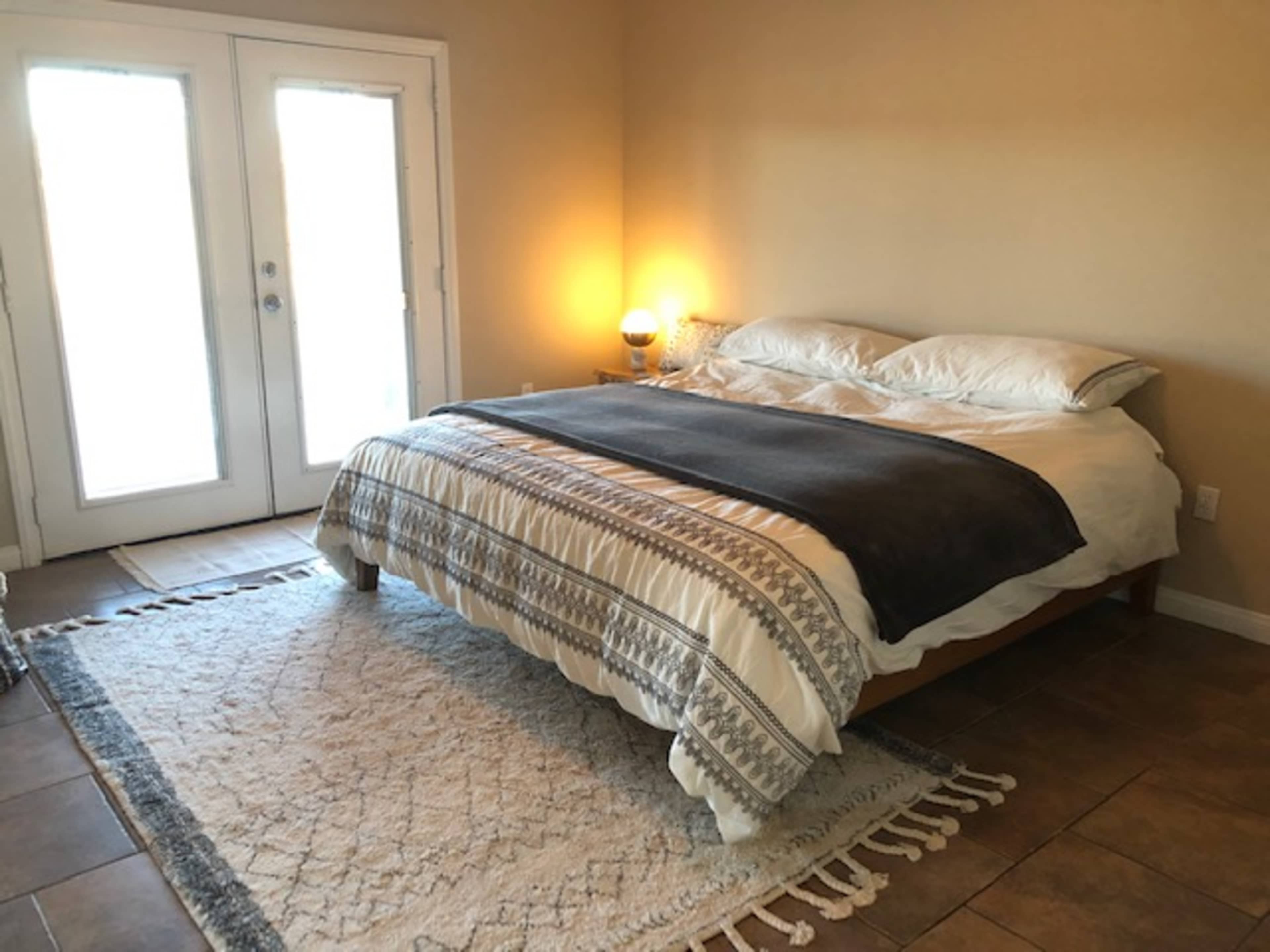 A bedroom featuring a bed with a patterned blanket, a soft rug on the floor, and two double doors leading to a bright outdoor area.