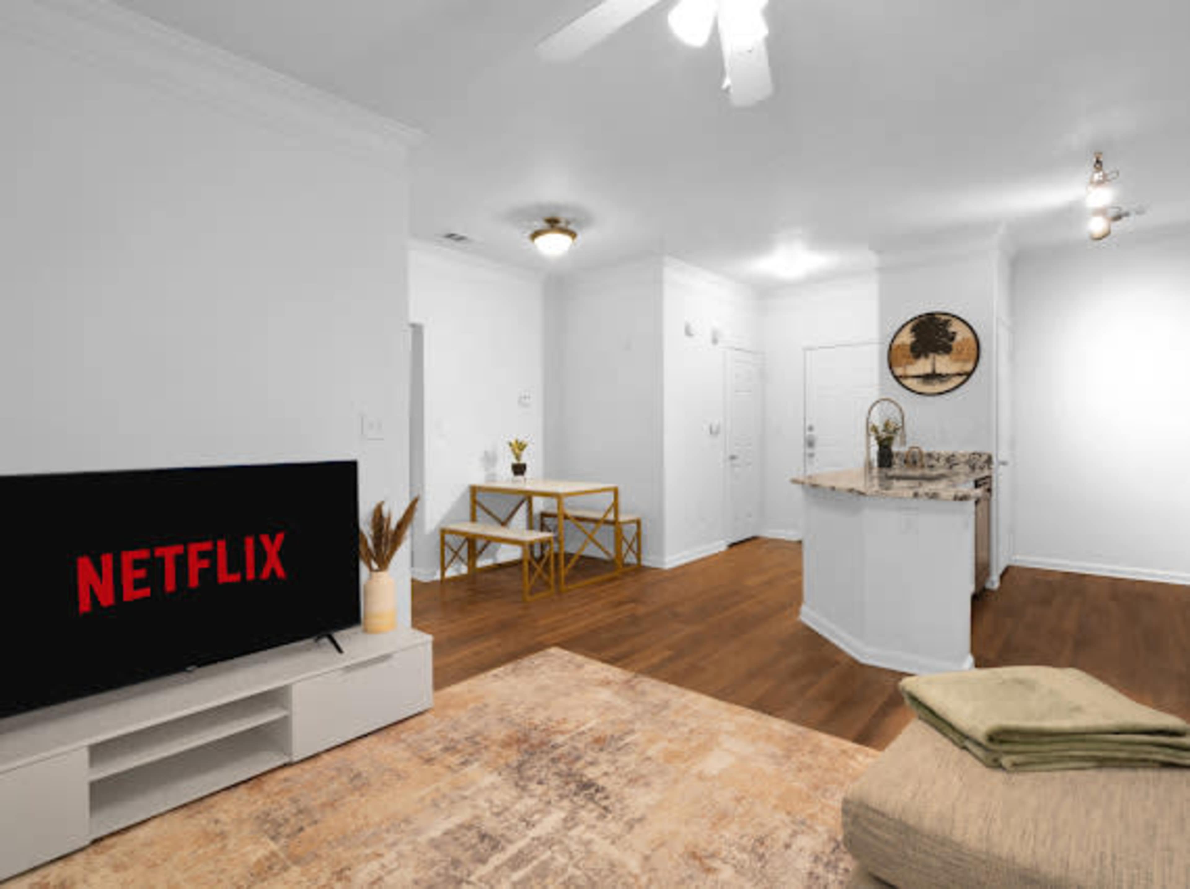 The image shows a modern living room with a large television displaying the Netflix logo, a dining area with a small table, and a kitchen in the background.