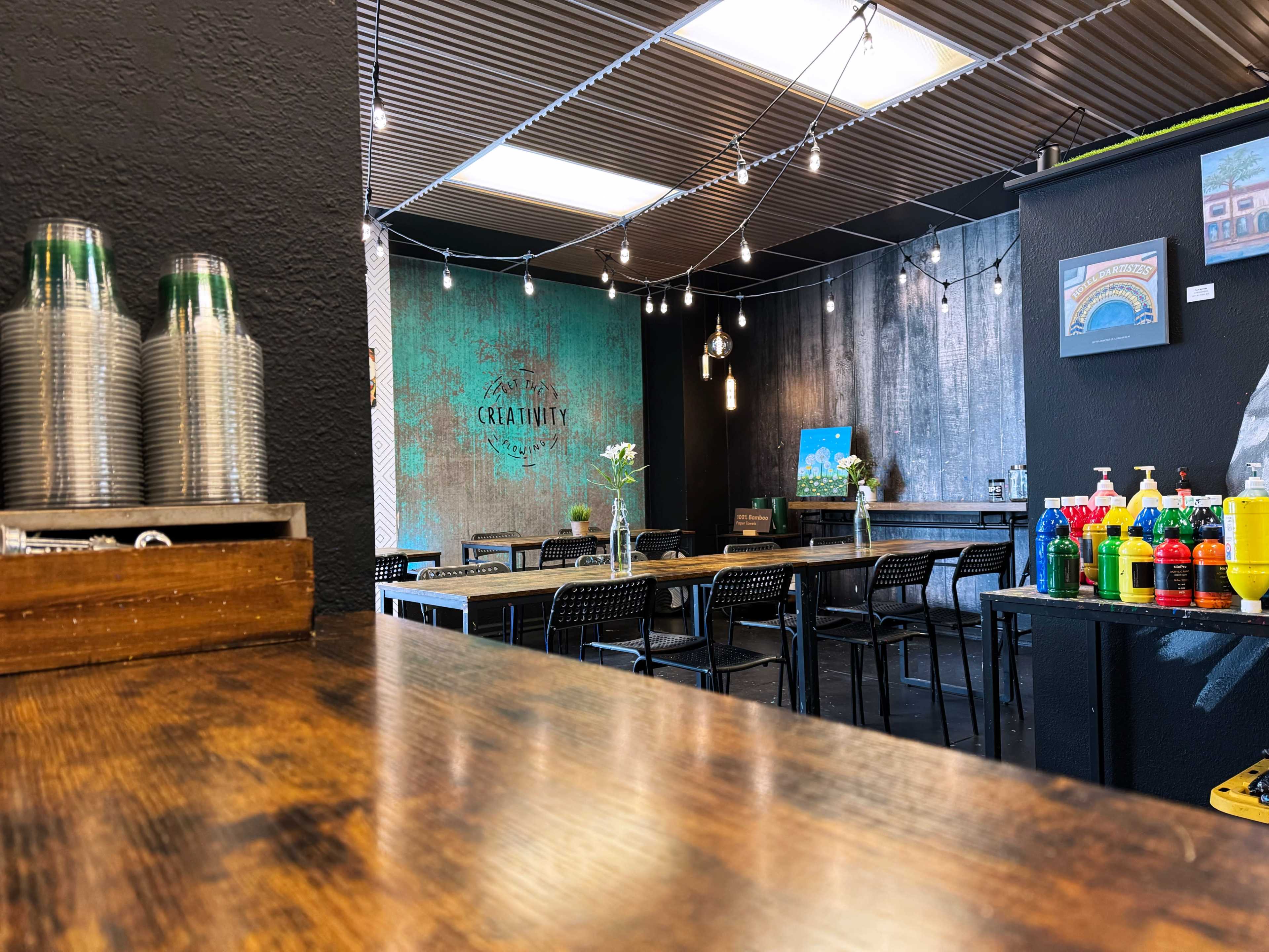 The image shows the interior of a restaurant with wooden tables, metal chairs, and decorative elements including hanging lights and colorful condiment dispensers.