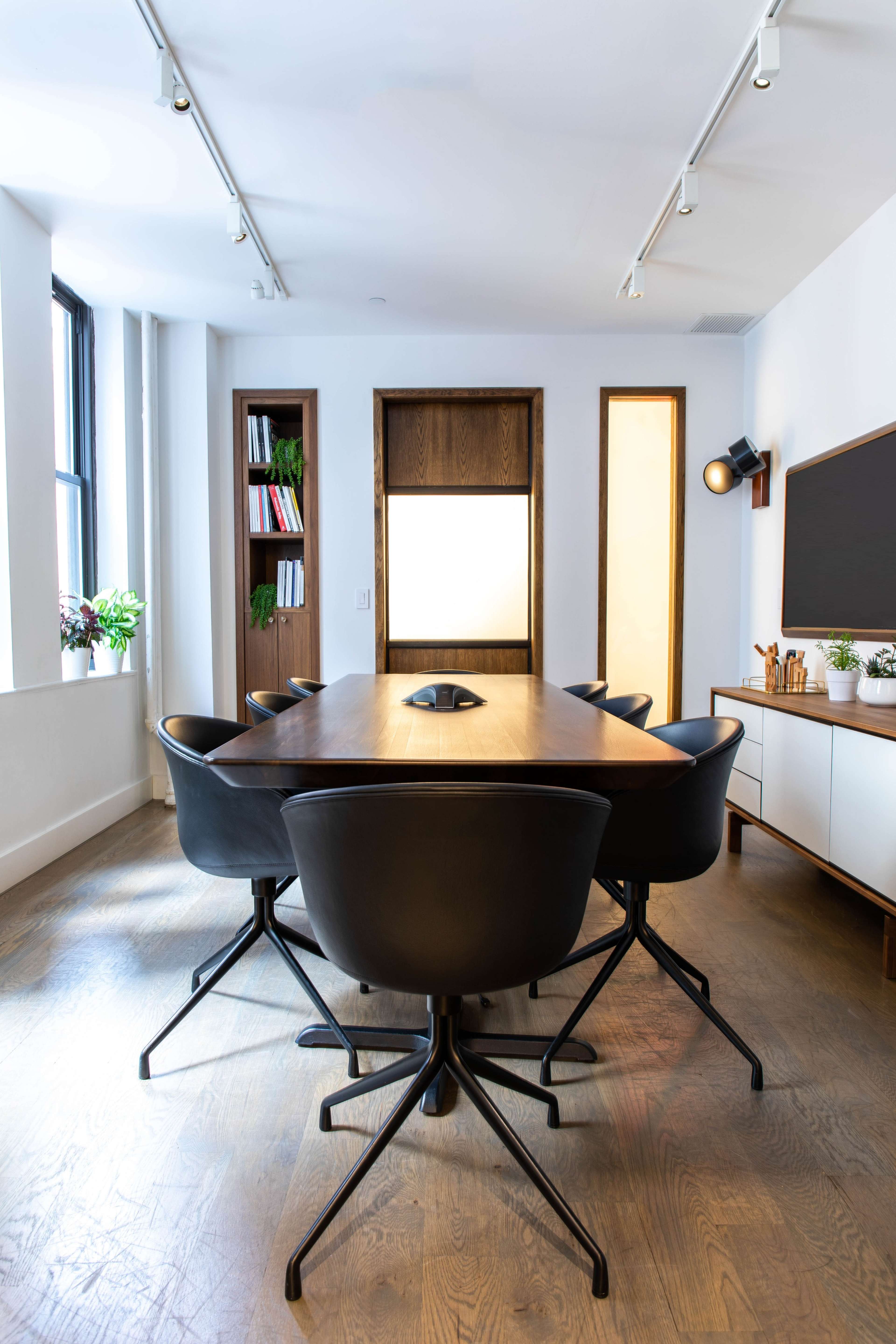 A modern conference room features a long wooden table surrounded by black swivel chairs, with large windows and built-in shelves displaying plants and books.