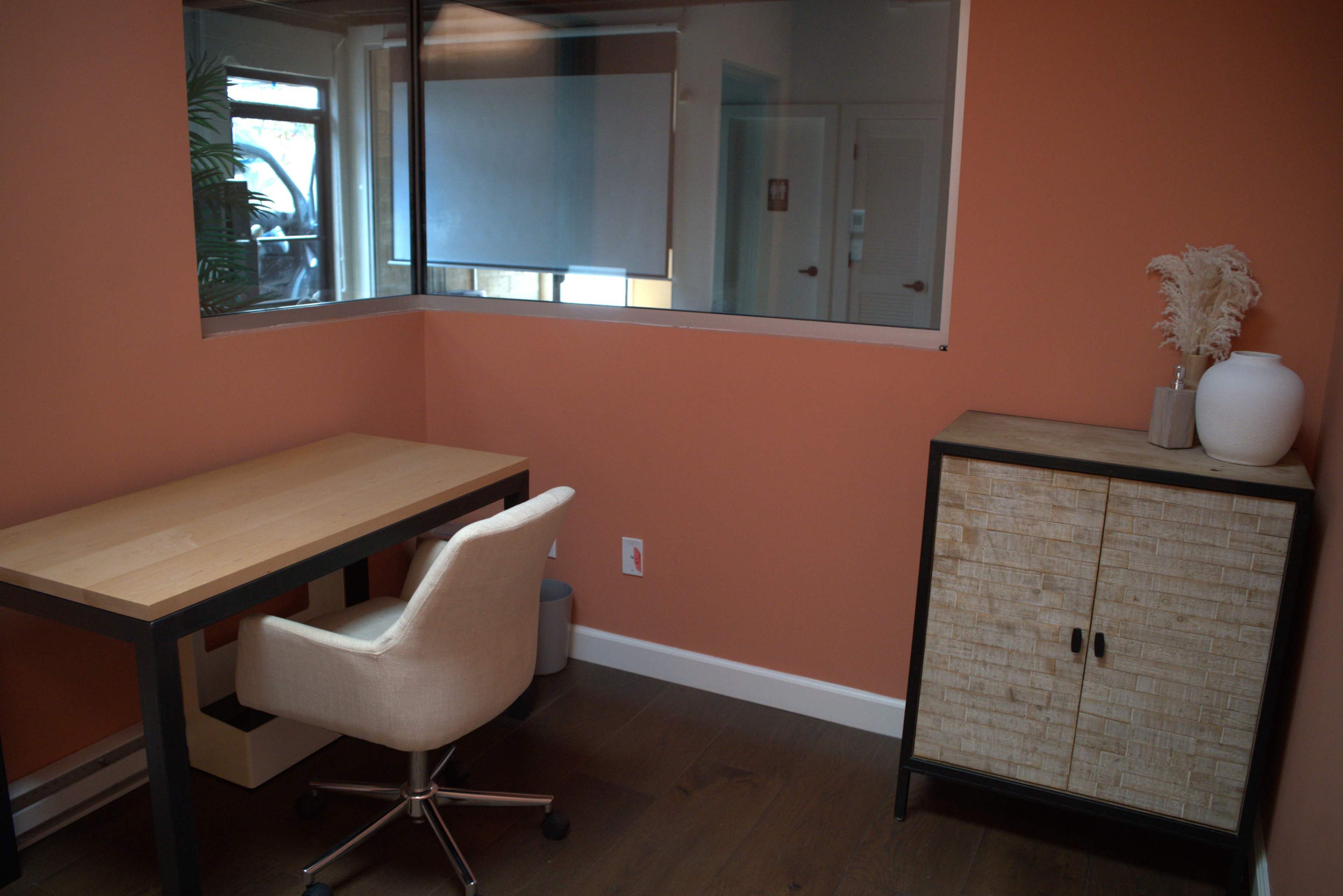 The image shows a small office space with a wooden desk, a white chair, and a storage cabinet against an orange wall.