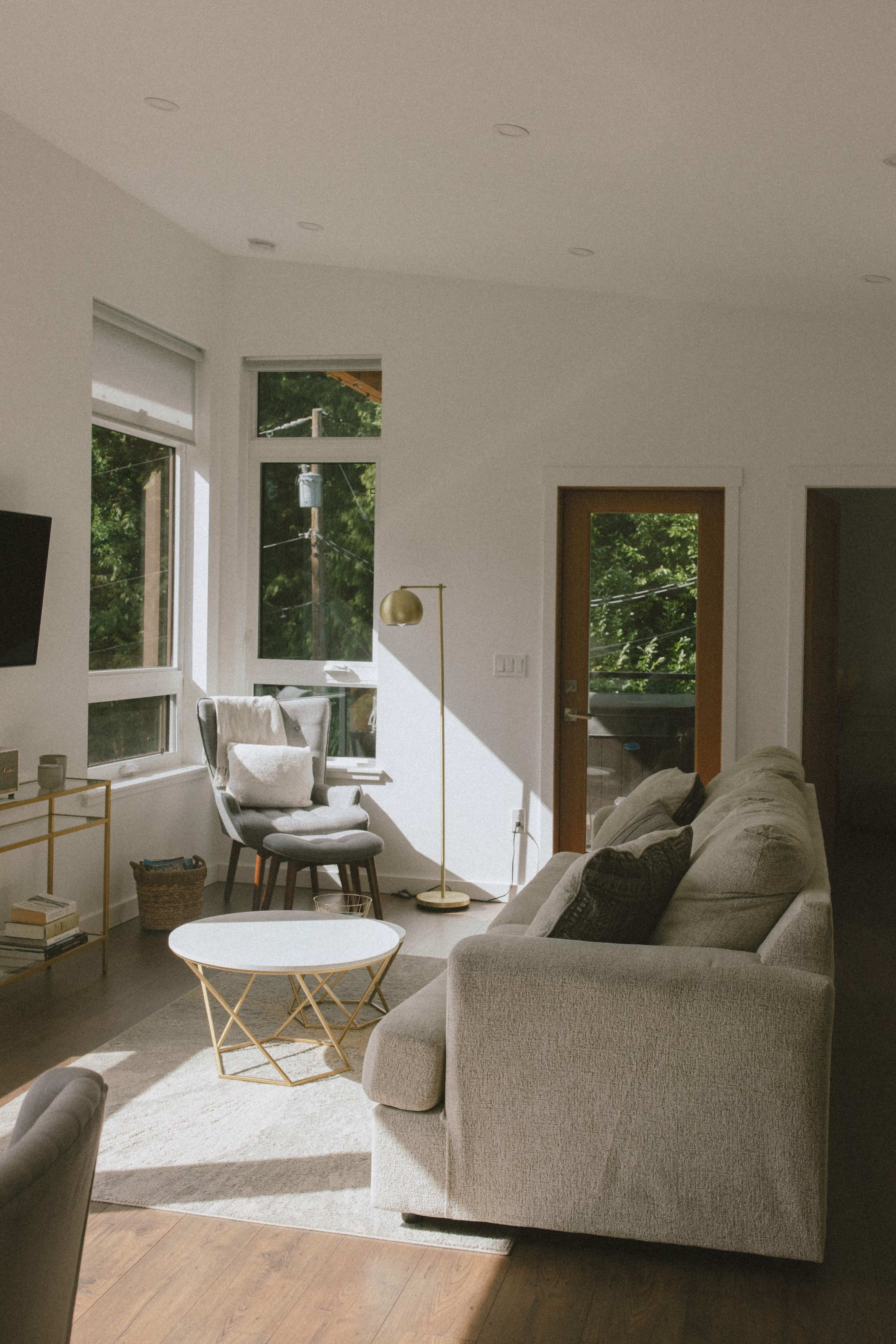 The scene shows a modern living room with large windows, a gray sofa, a round coffee table, and a floor lamp beside an armchair, leading to a door that opens to the outside.