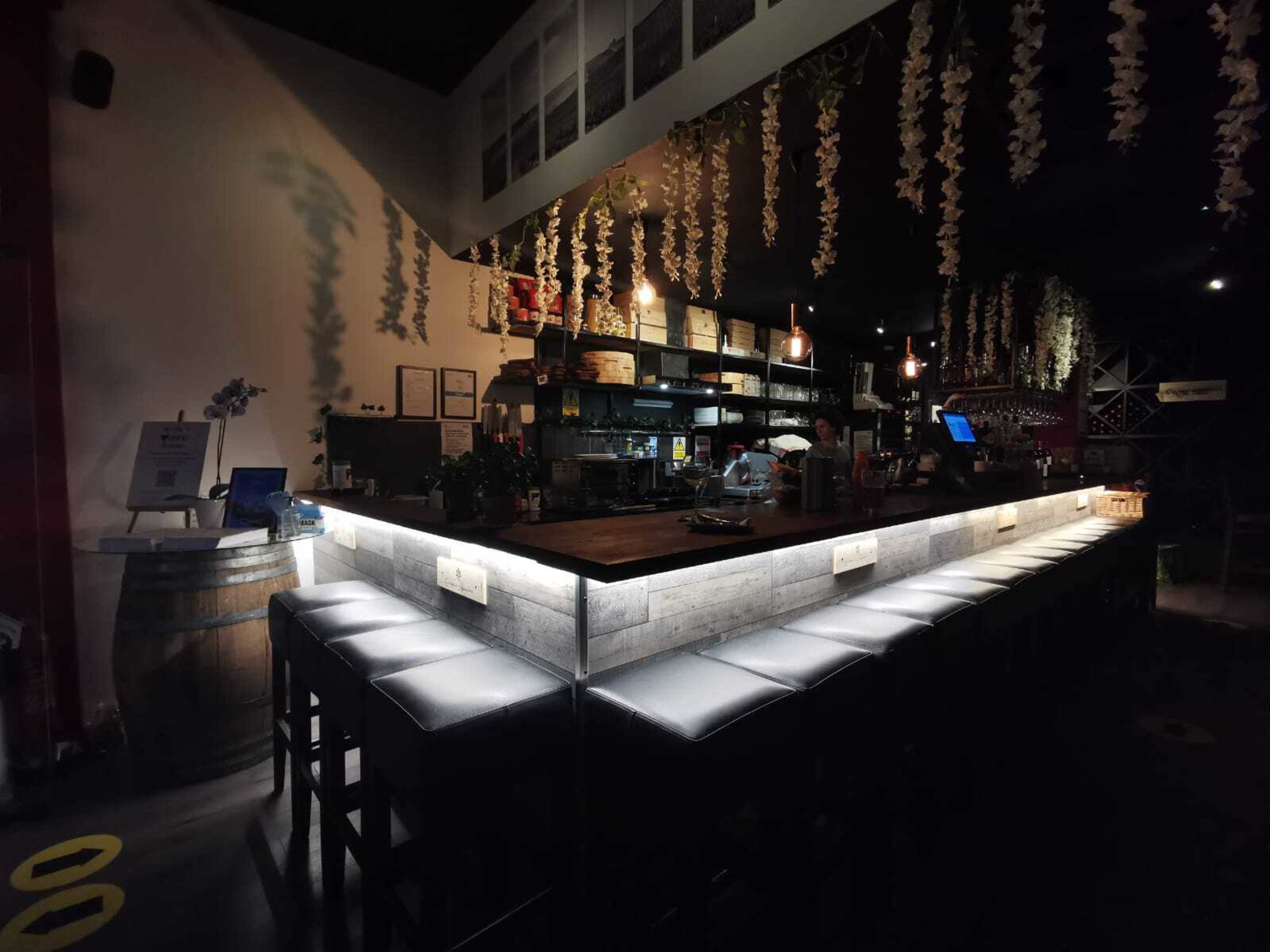 A dimly lit bar features a long, illuminated counter with high stools and shelves lined with various bottles.