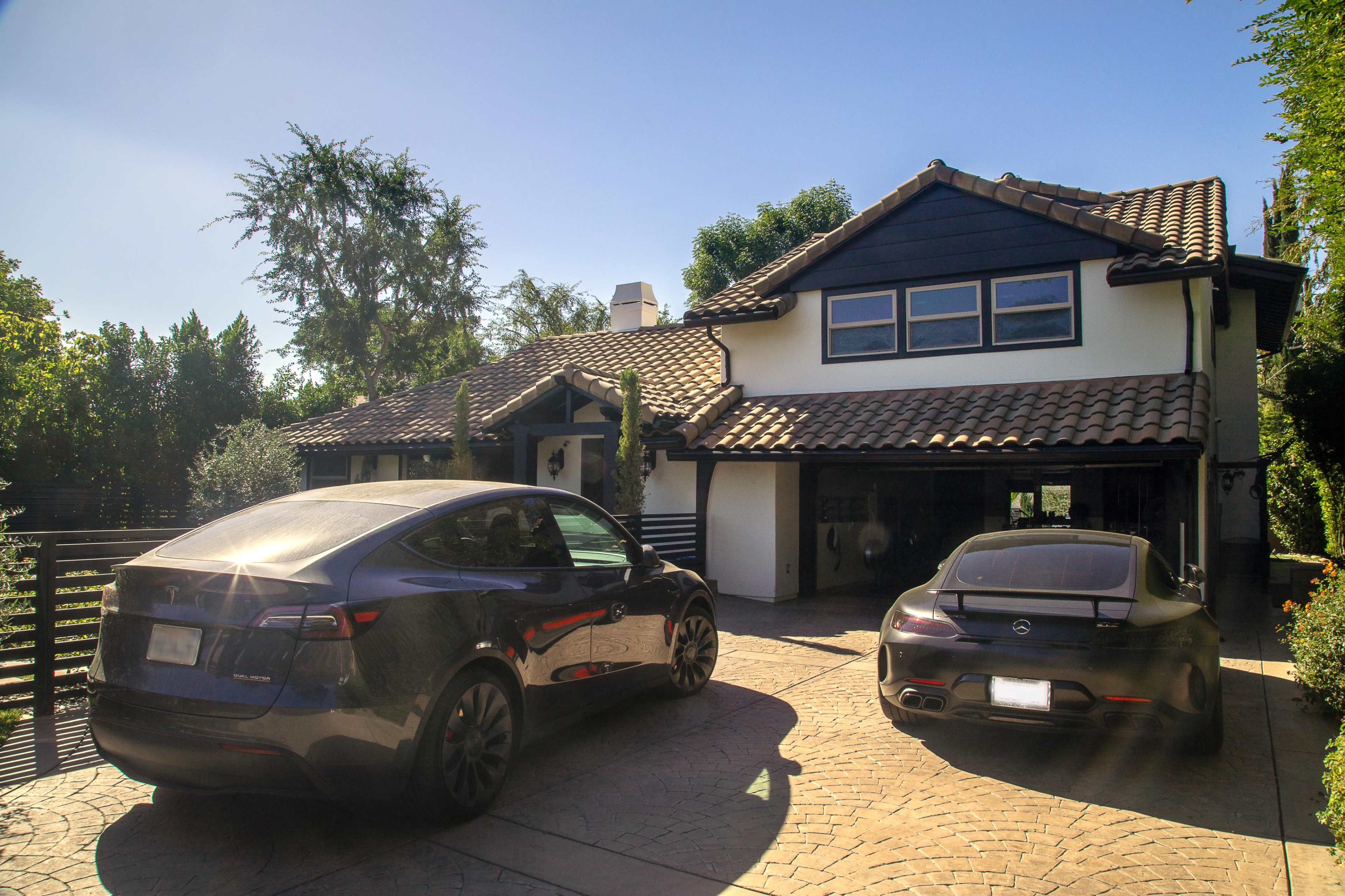Two cars, a Tesla Model Y and a Mercedes-Benz, are parked in the driveway of a two-story house with a tiled roof and lush landscaping.