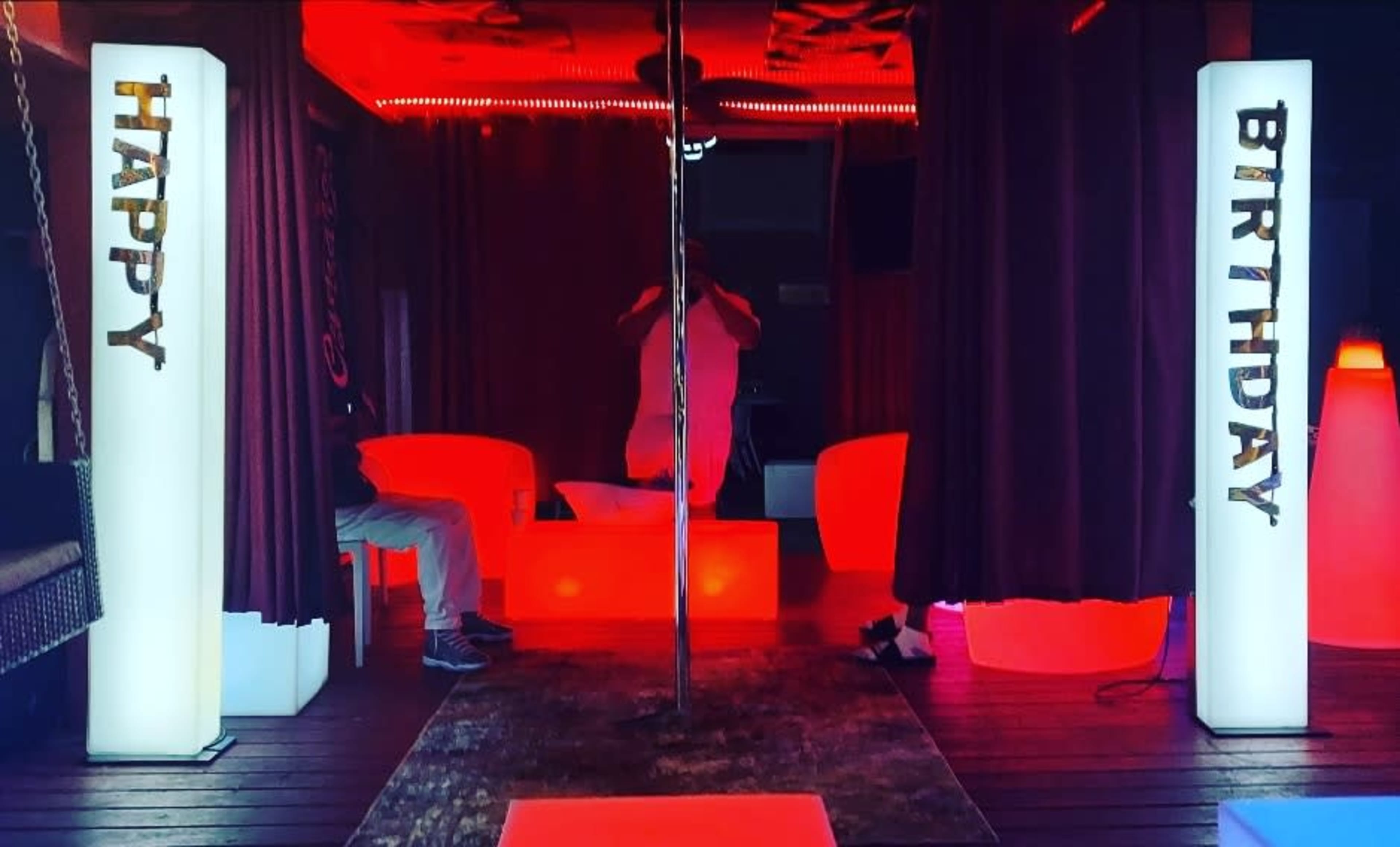 The image shows a party space decorated with glowing "HAPPY" and "BIRTHDAY" signs illuminated in red light, alongside a pole in the center.