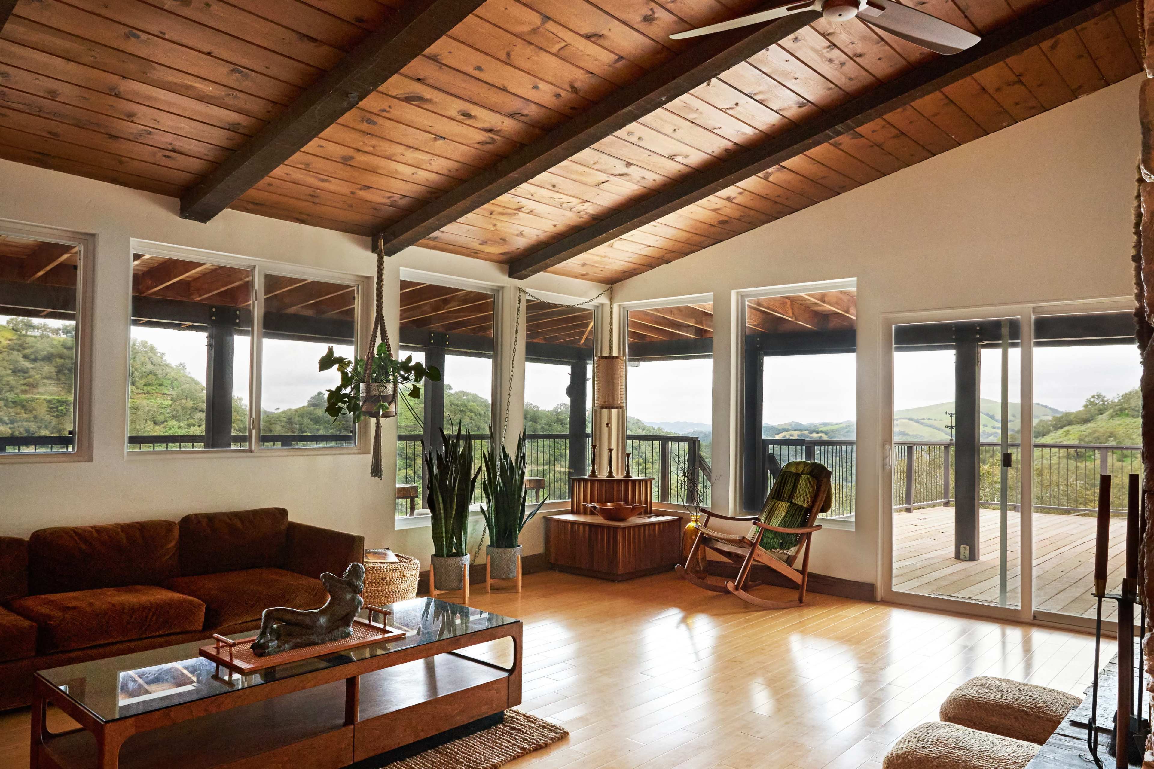The interior of a modern living room features a brown sofa, a glass coffee table, and large windows that provide a view of the outdoor landscape.