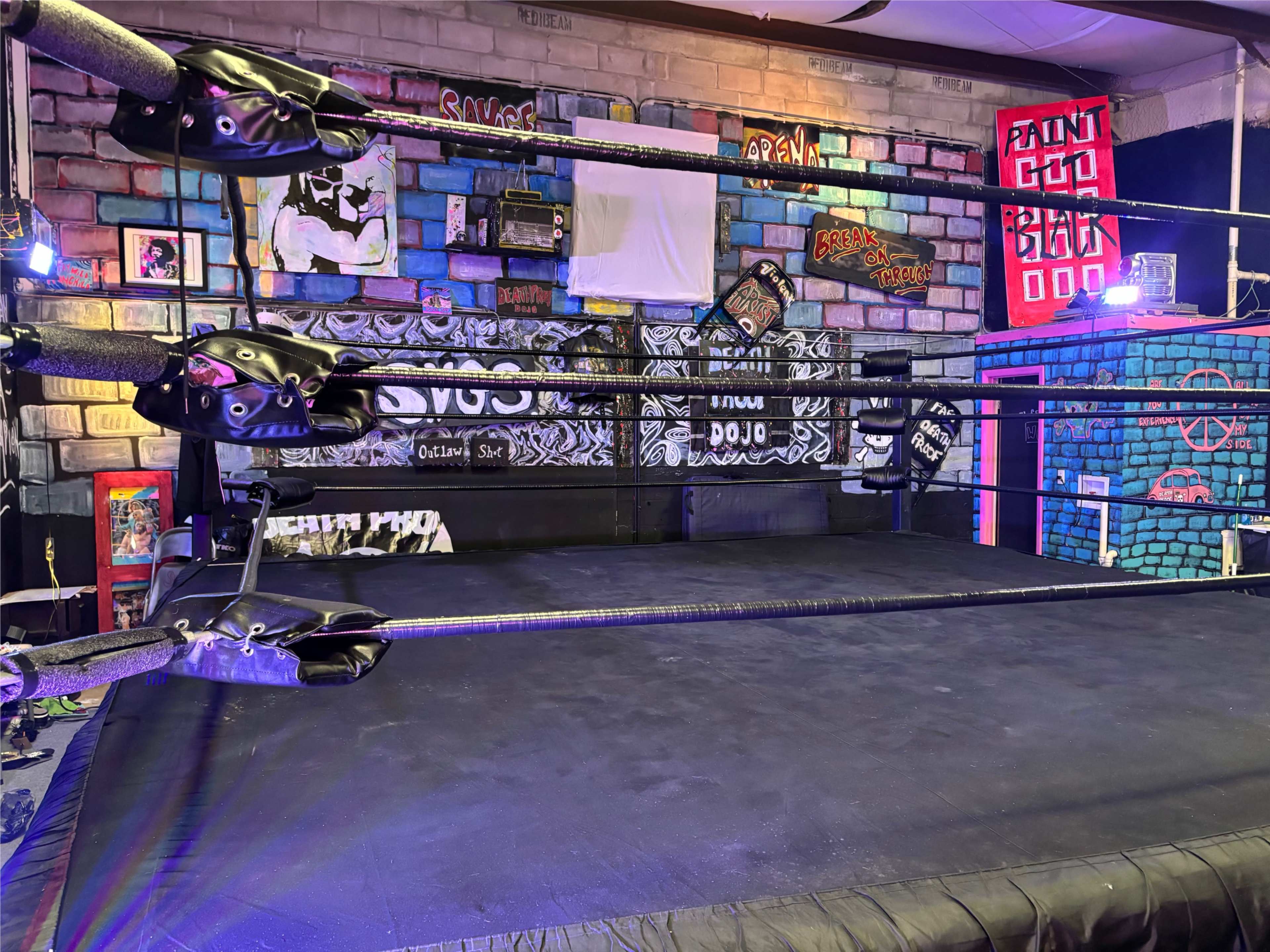 The image shows a wrestling ring set up in a colorful indoor space with graffiti-covered walls and various art pieces.