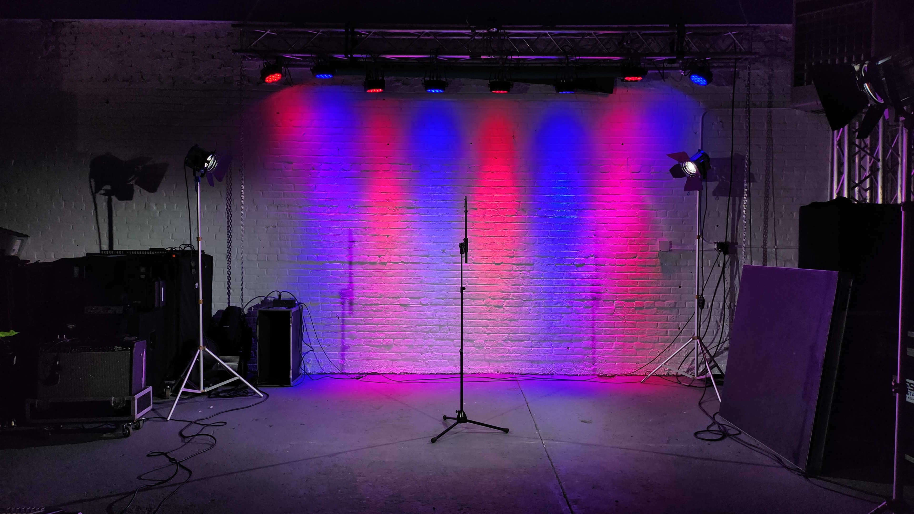A performance stage with colored lights illuminating a microphone stand in front of a lit white brick wall.
