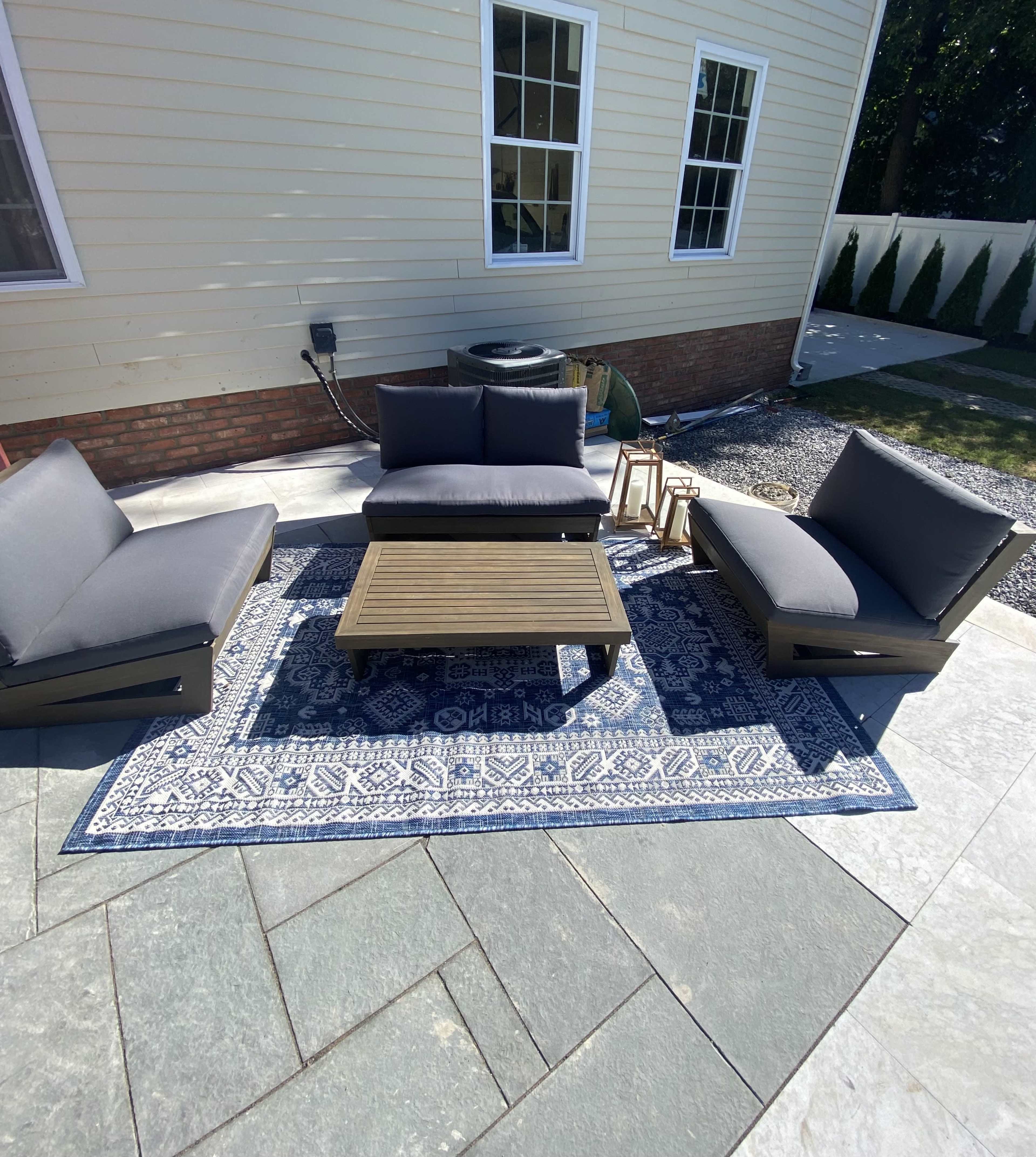 A patio seating arrangement with two chairs, a sofa, and a coffee table positioned on a patterned area rug, set against a stone floor and a house with large windows.