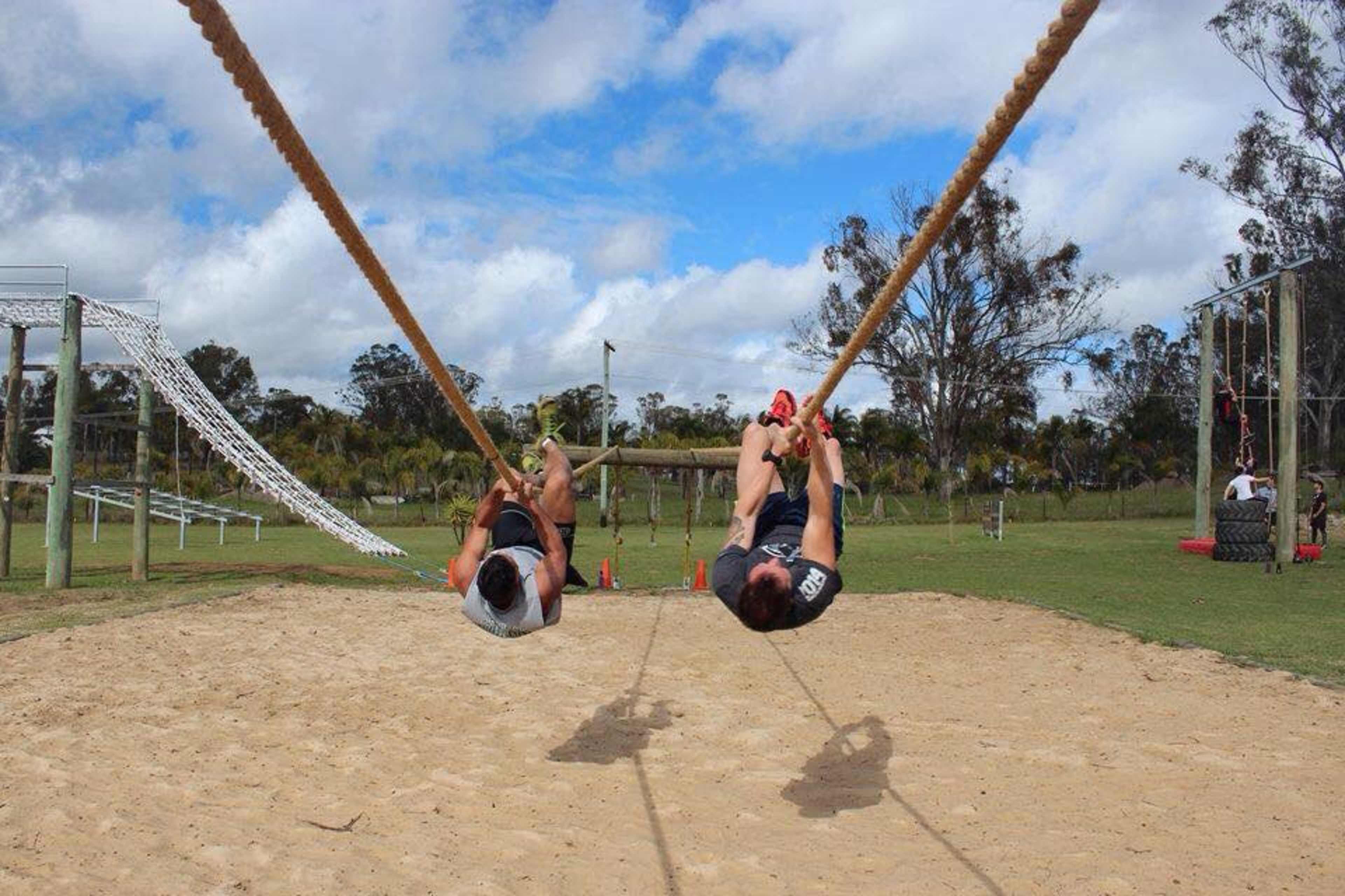 Two men are swinging on ropes above a sandy area in a playground, with equipment and trees in the background.