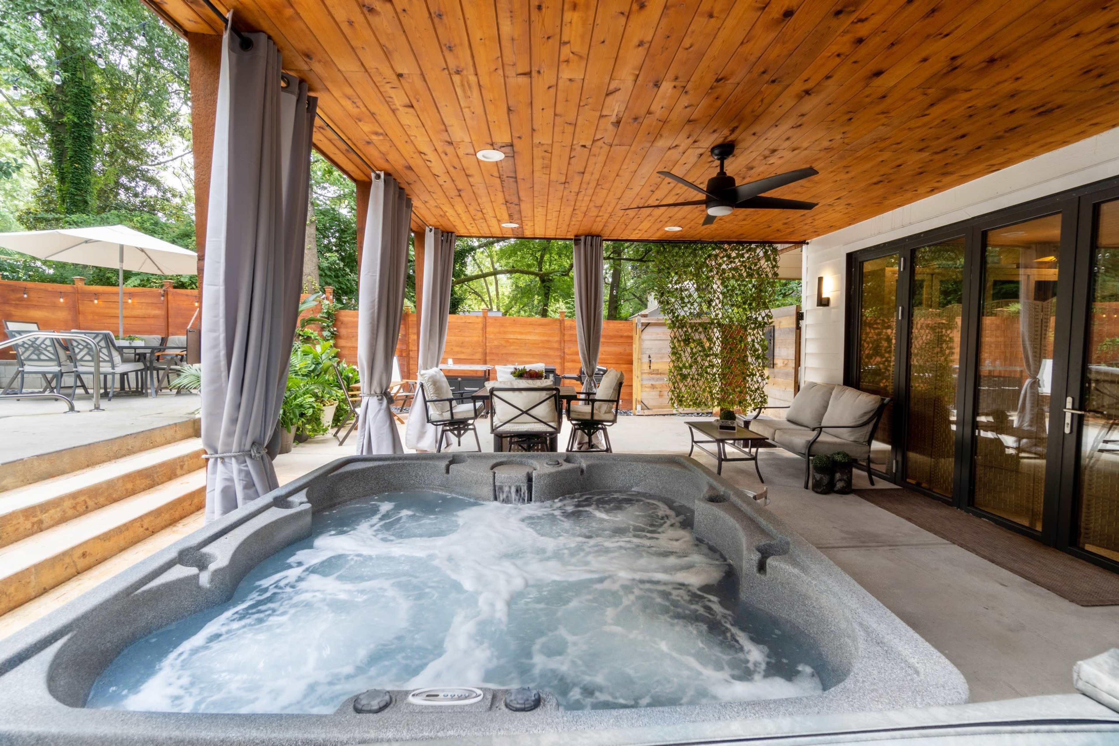 A covered outdoor patio featuring a hot tub, dining area, and seating arrangements surrounded by greenery.