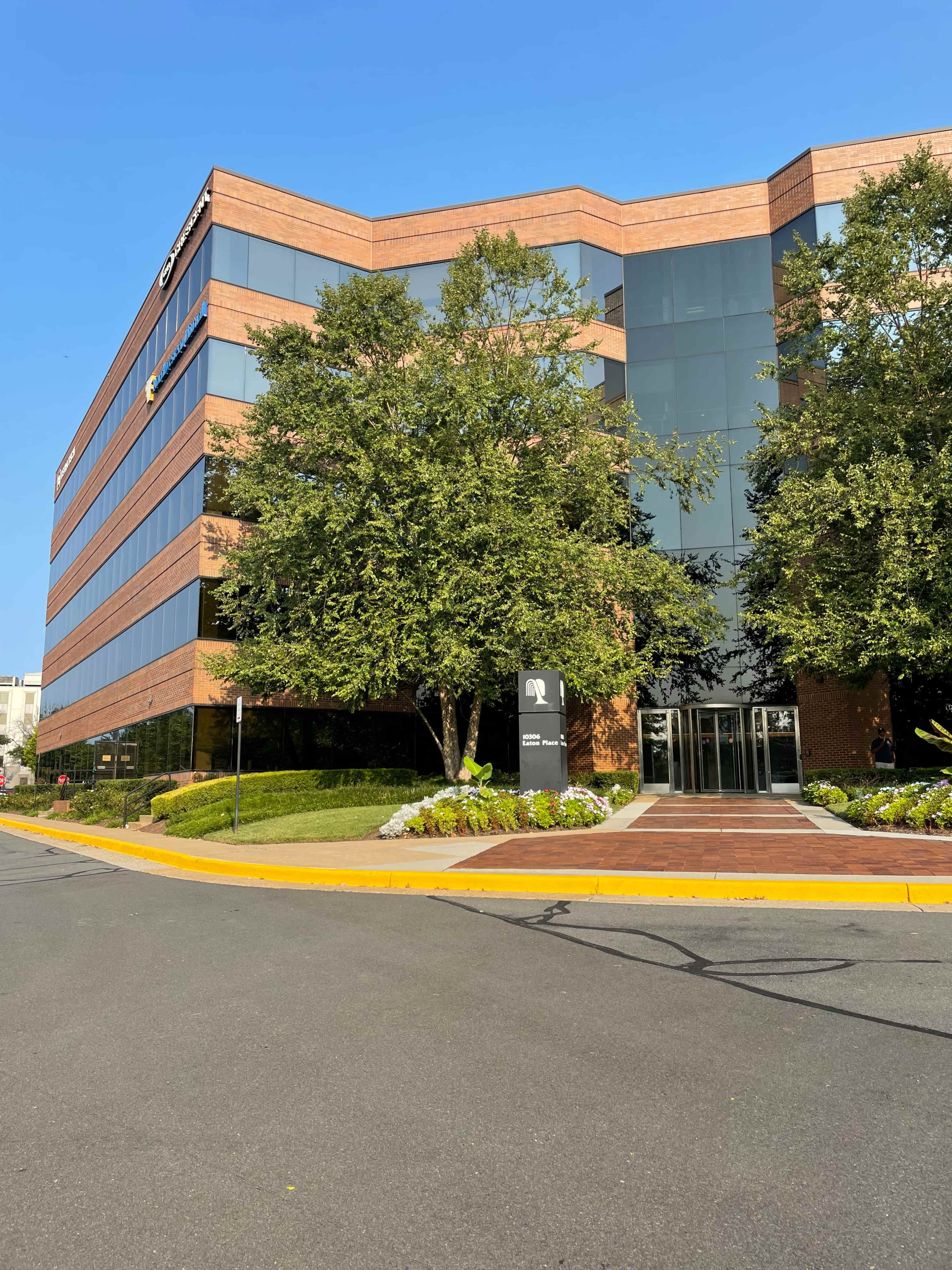 A modern brick office building with glass windows is surrounded by green landscaping and a paved entrance.