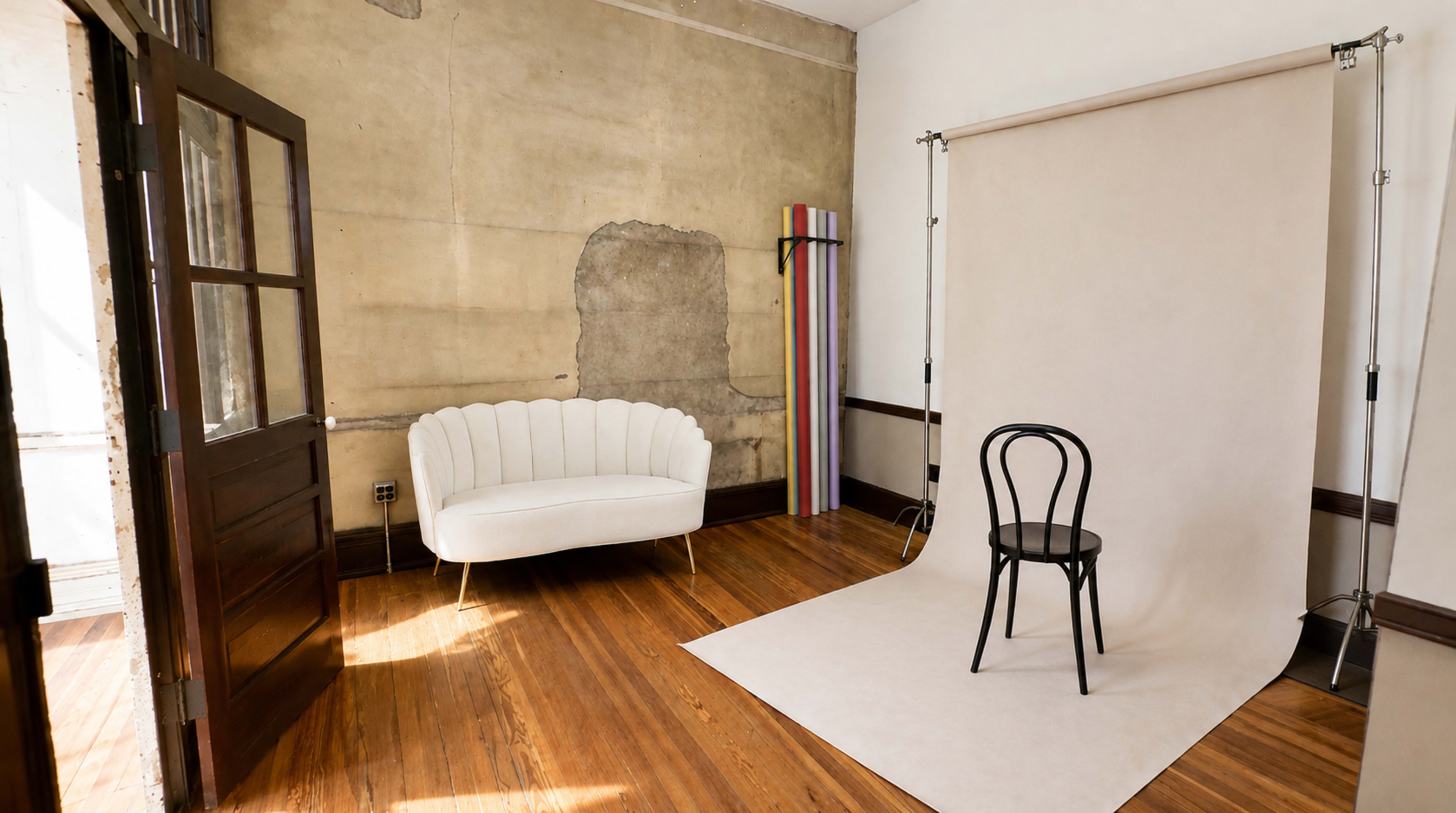 The image shows an empty photography studio with a white backdrop, a black chair in front of it, and a white sofa against a textured wall.