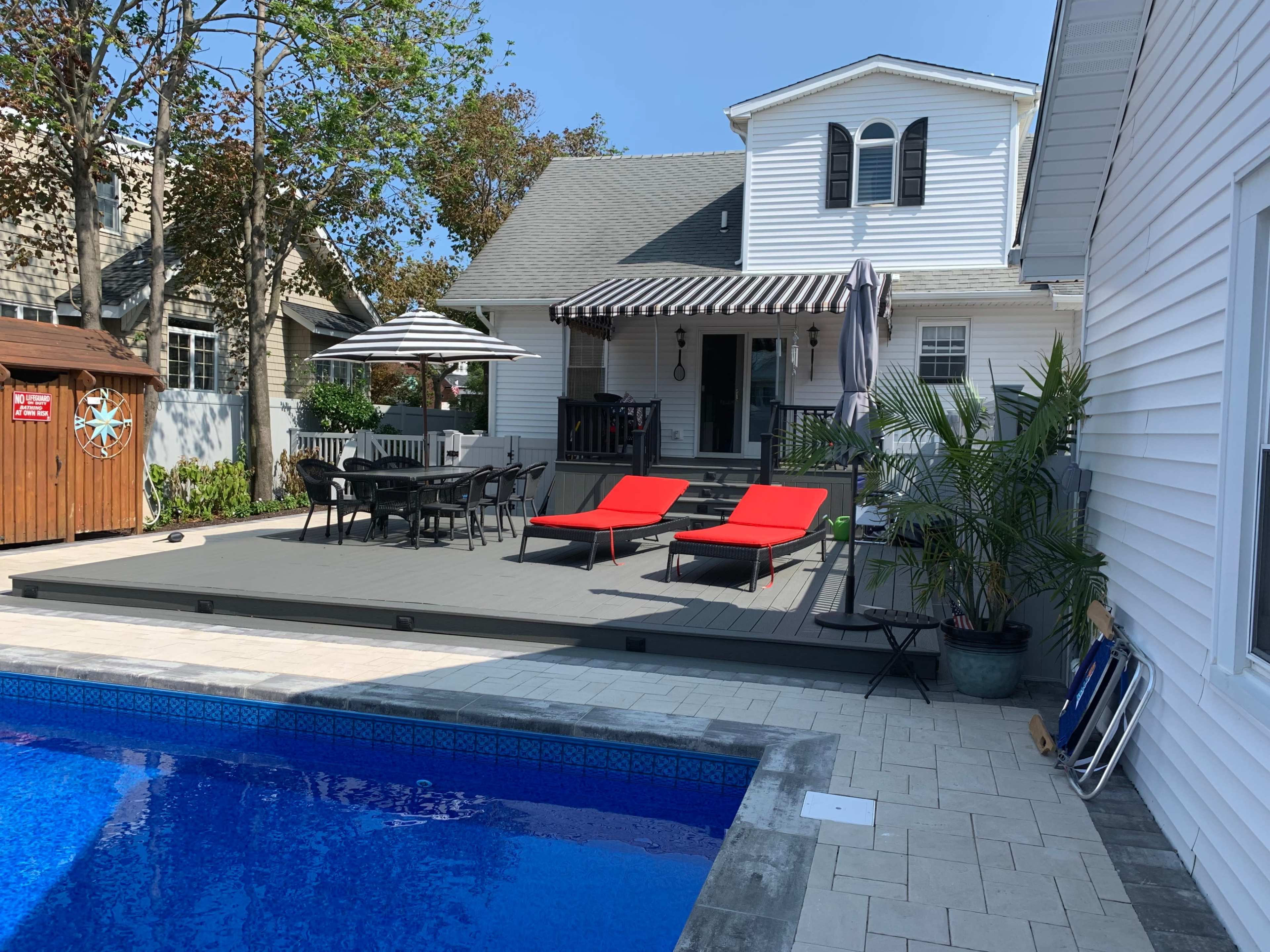 A backyard scene features a swimming pool surrounded by a deck with red lounge chairs, a dining area under an umbrella, and a house with white siding in the background.