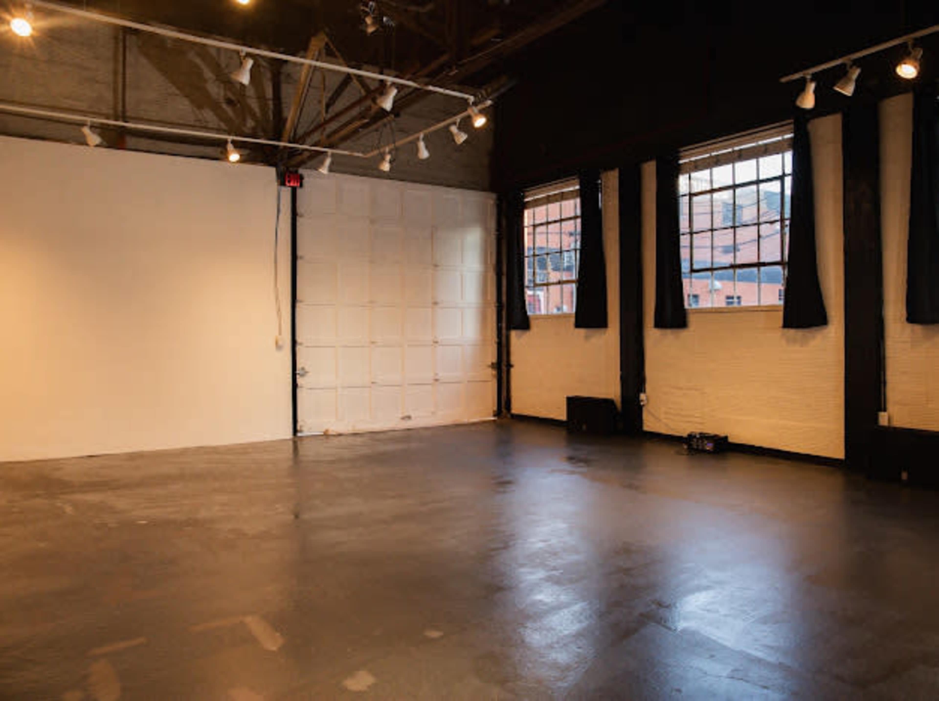 The image shows a spacious, empty room with large windows and exposed ceiling beams, featuring a white wall on one side and dark curtains on the windows.