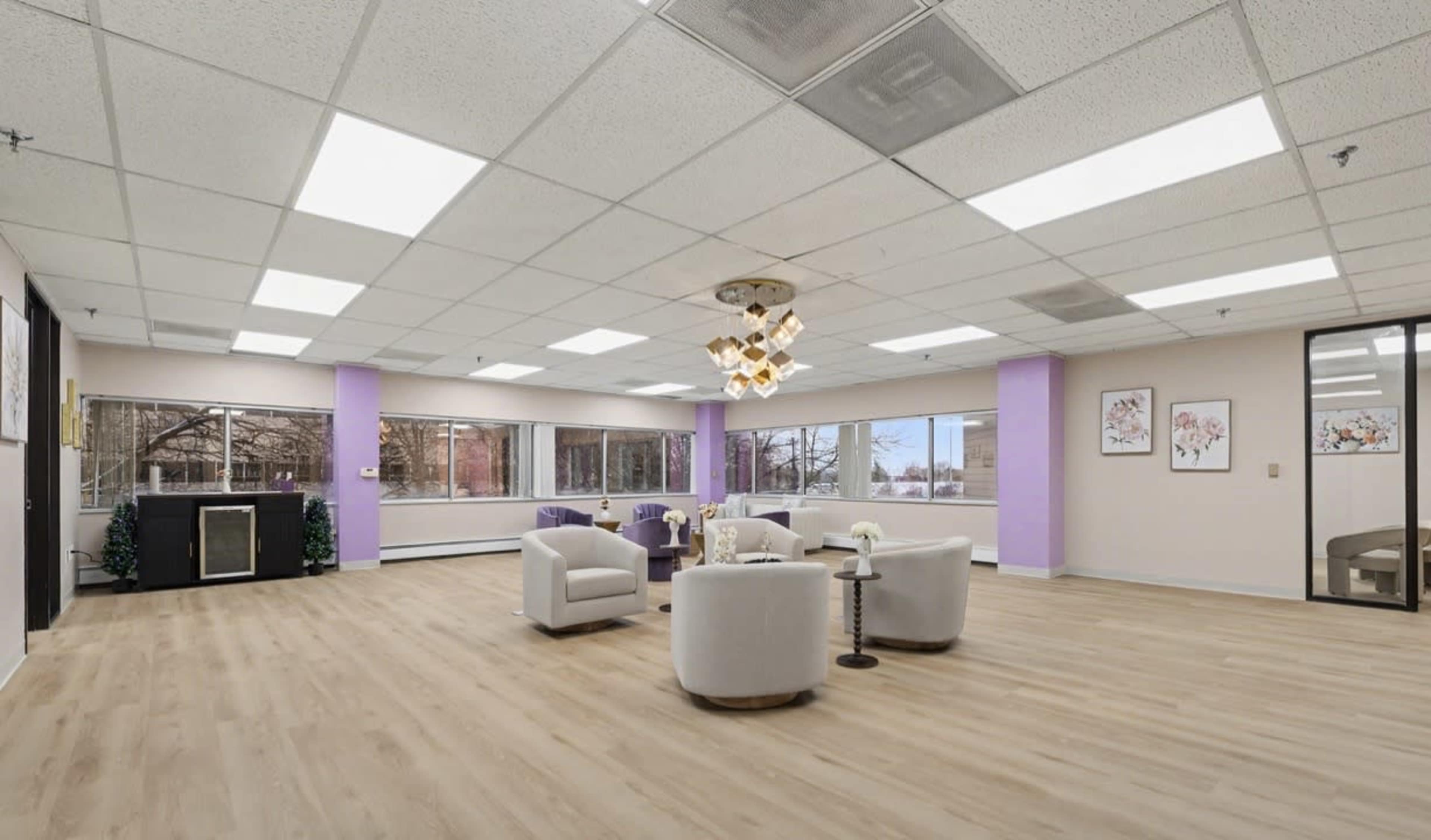 The spacious, brightly lit room features a circular seating arrangement with white upholstered chairs and a modern chandelier, surrounded by large windows.