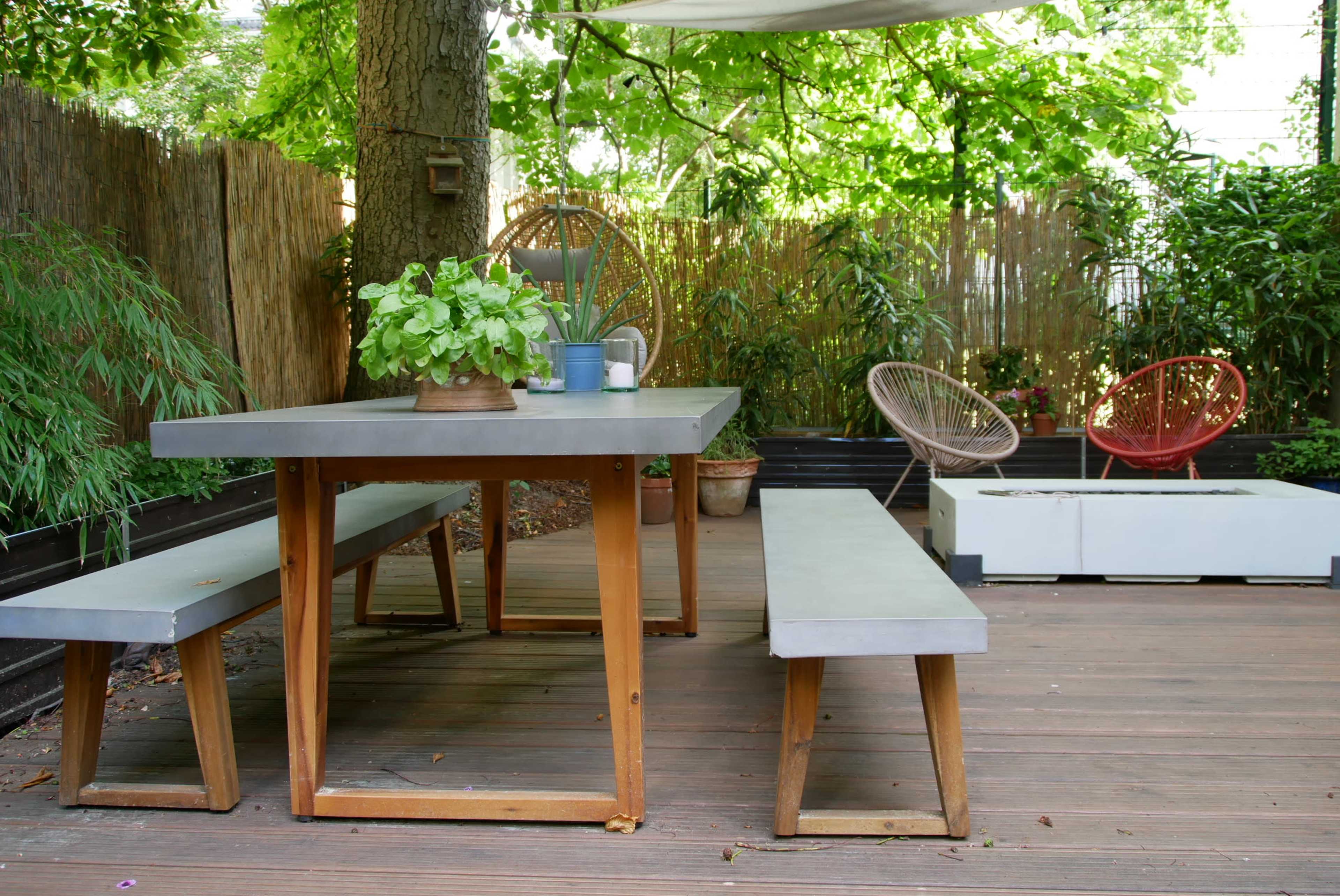 A wooden outdoor dining table with benches is surrounded by greenery in a shaded patio area.