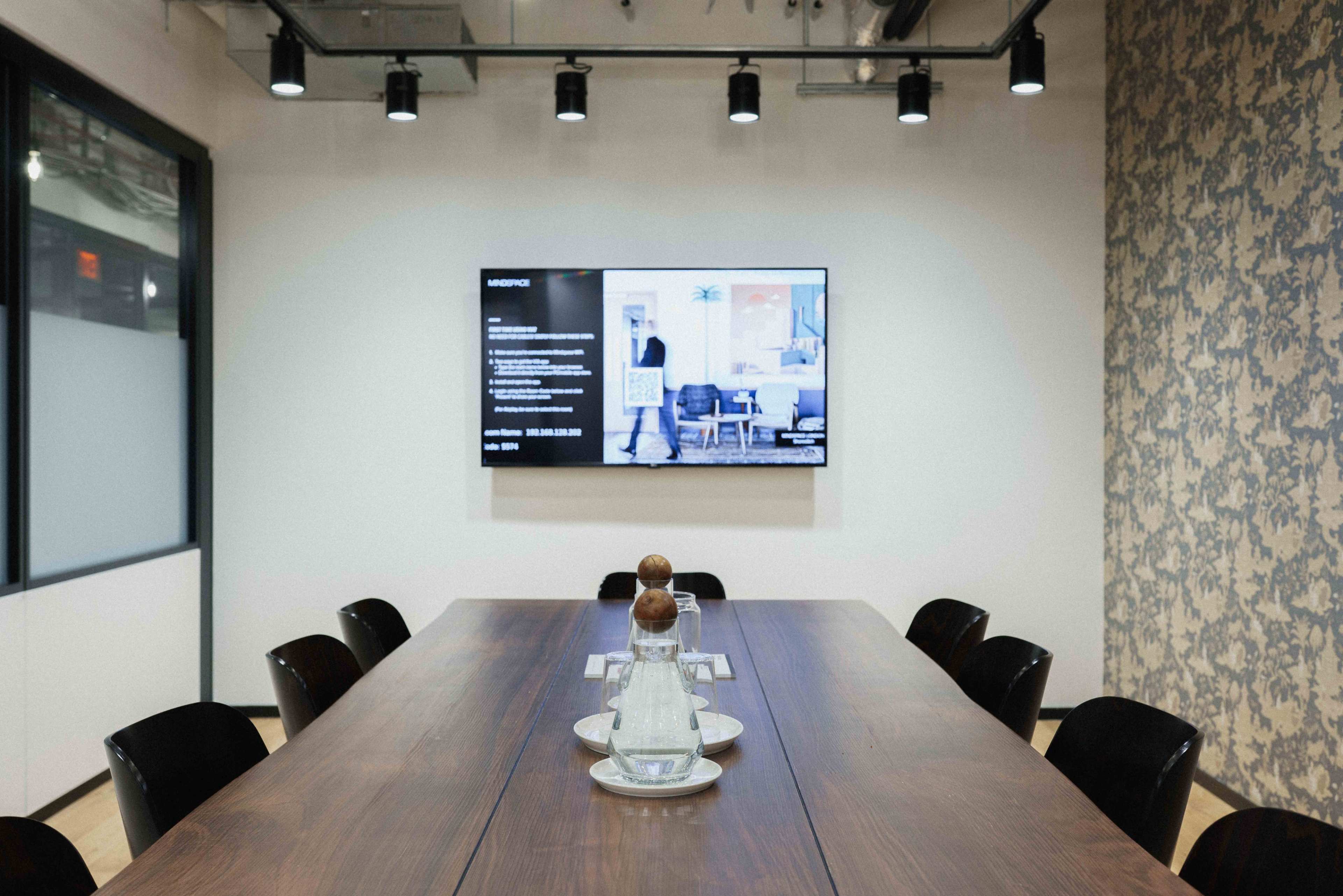 A conference room features a long wooden table, a large wall-mounted screen displaying information, and black chairs arranged around the table.