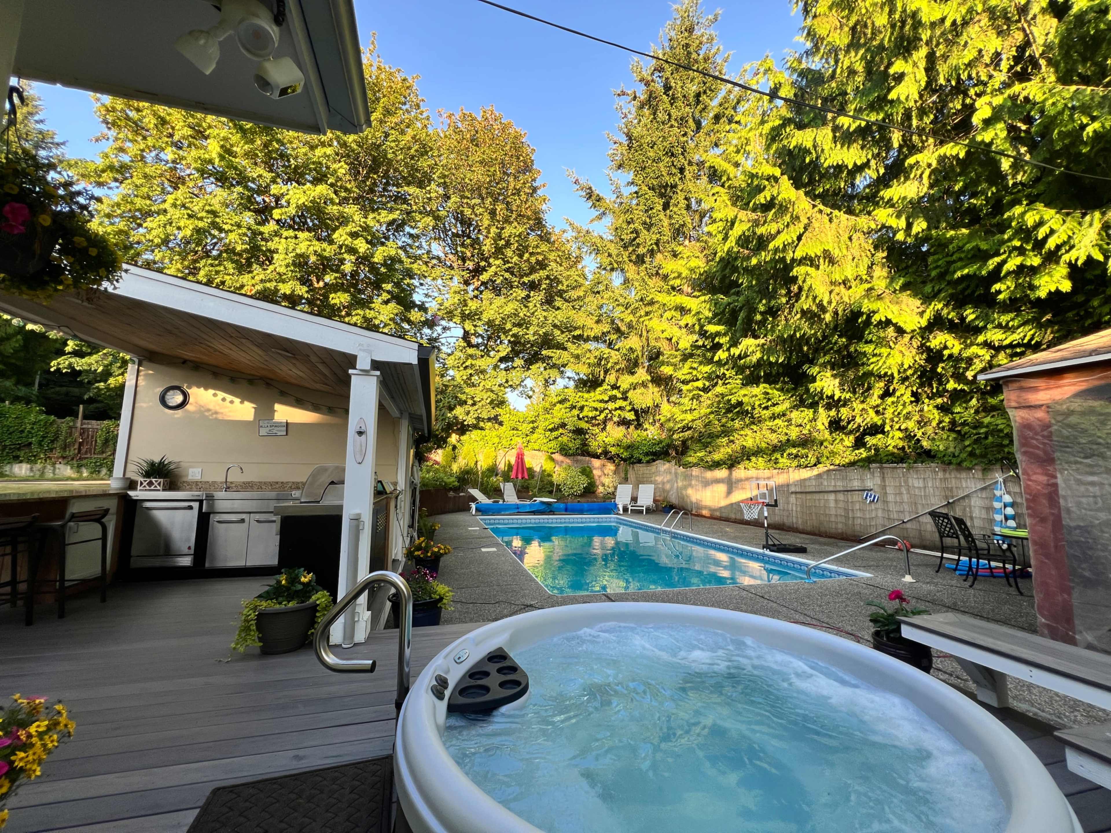 The image shows an outdoor area featuring a hot tub, a swimming pool, and a surrounding deck with trees in the background.