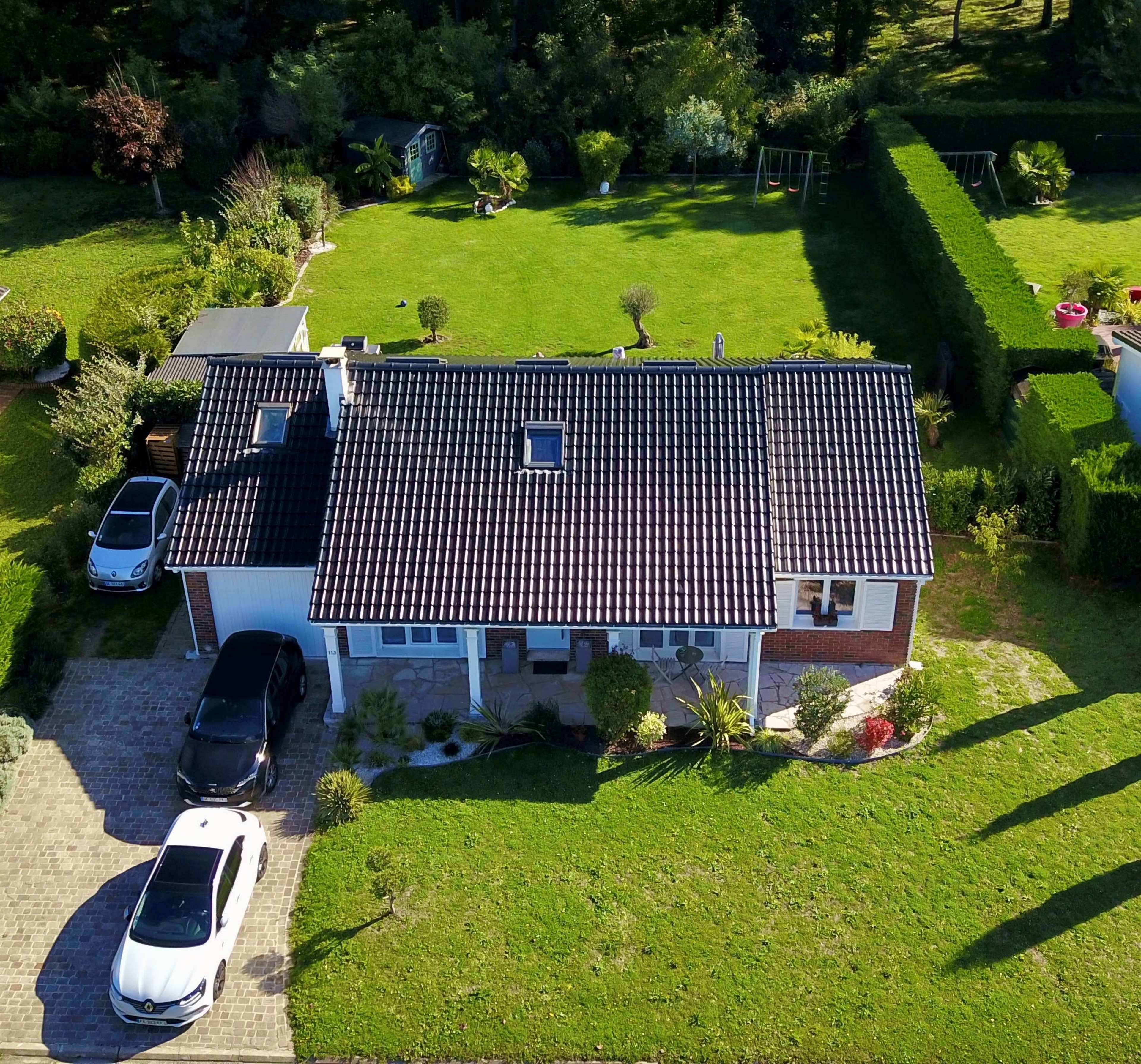 An aerial view shows a suburban house with a tiled roof, surrounded by a green lawn and landscaping.