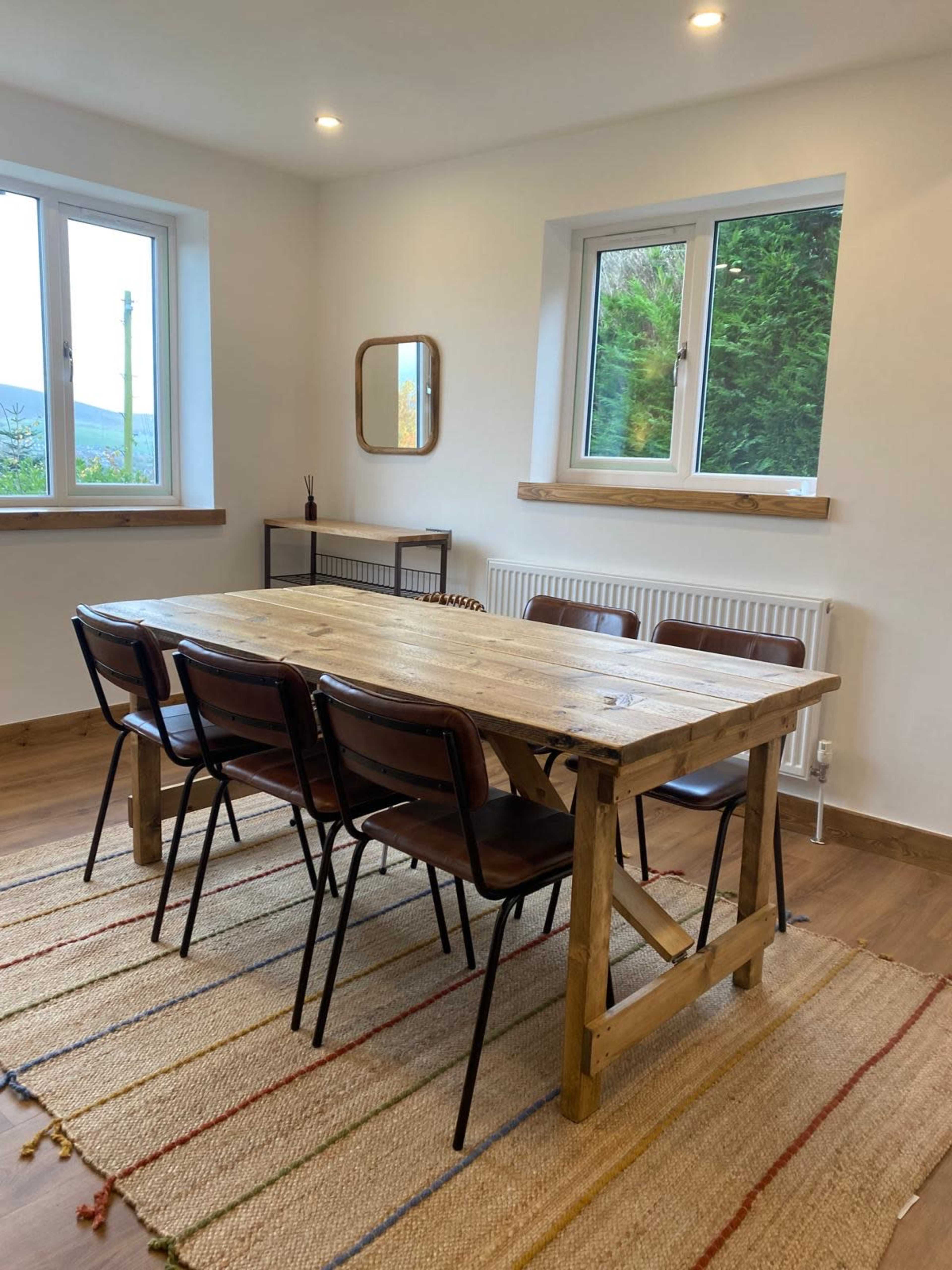 A dining area features a wooden table surrounded by six metal chairs, with large windows allowing natural light and a view of greenery.