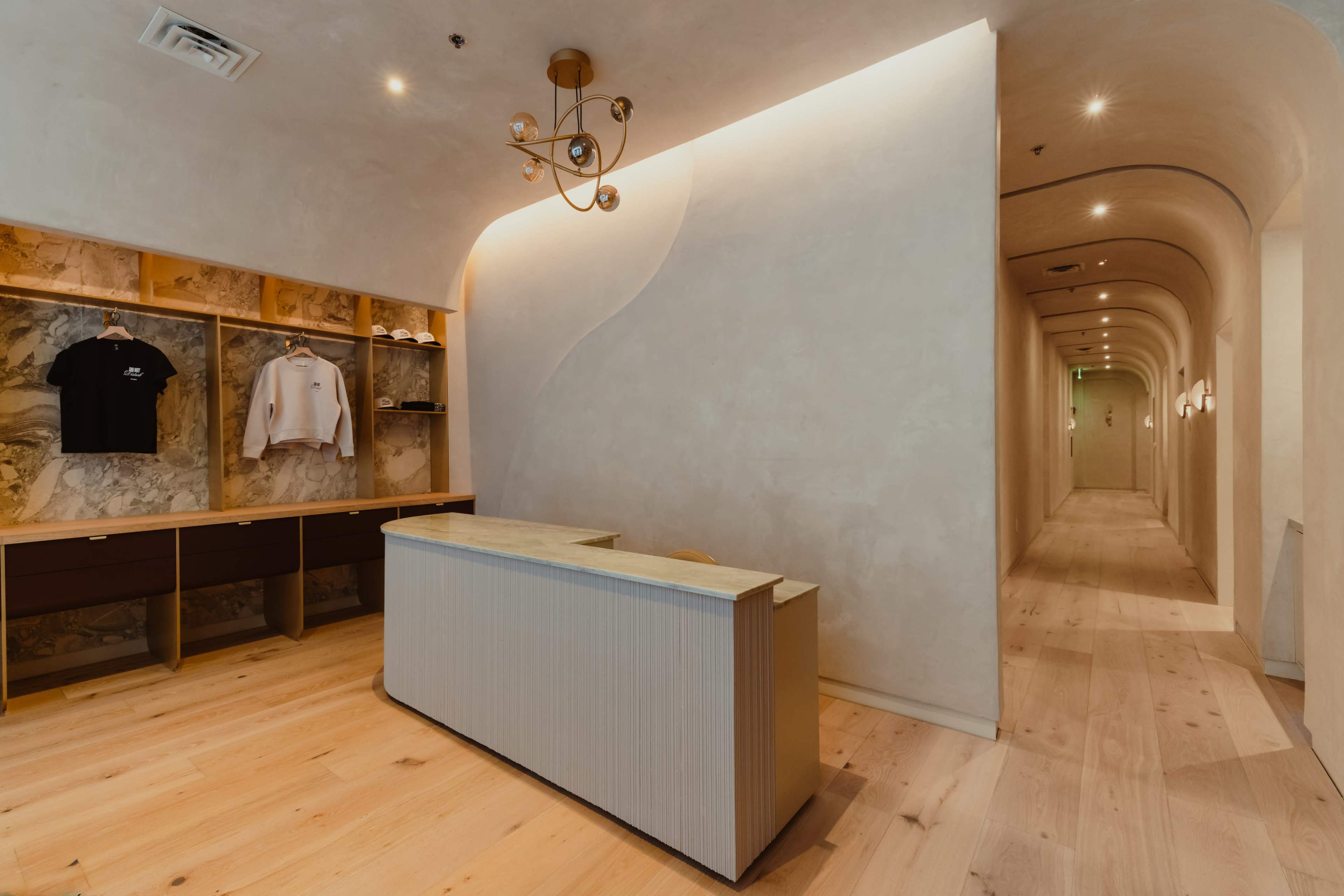 The image shows a minimalist reception area with a wooden desk and decorative wall shelves, leading to a narrow hallway with soft lighting and smooth walls.