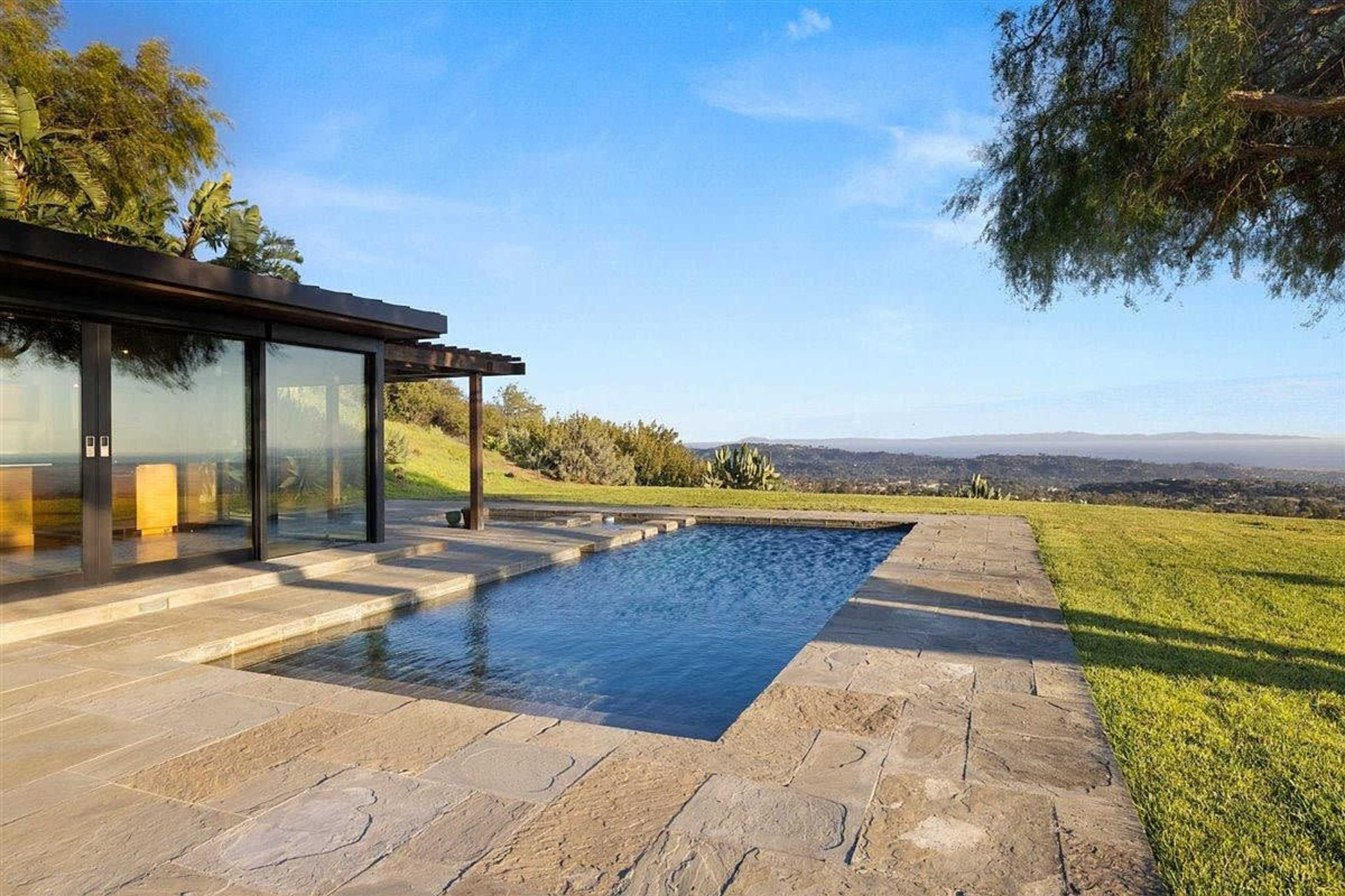 A modern house with large glass windows overlooks a swimming pool and a sprawling landscape.