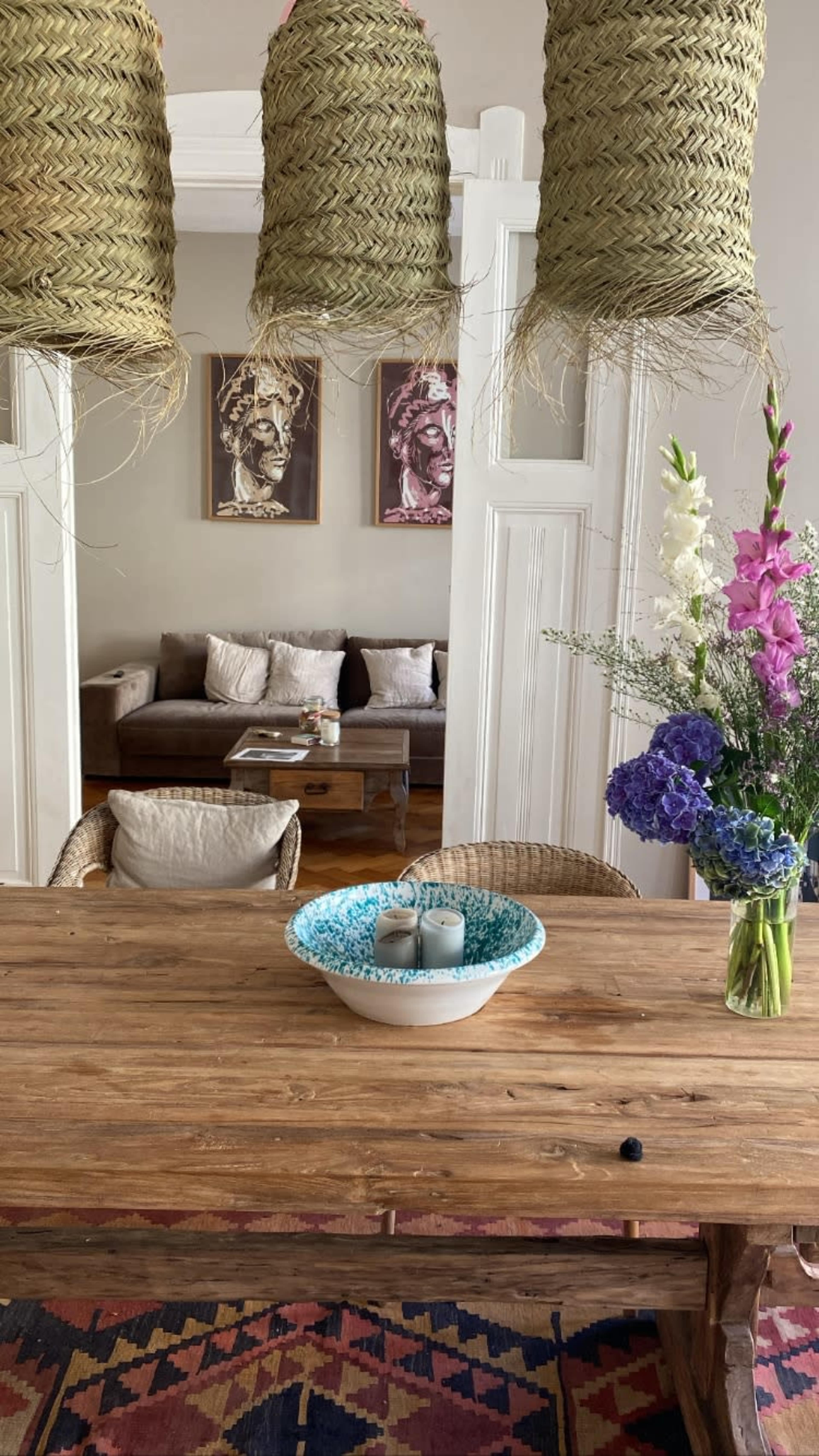 A wooden dining table with a decorative bowl and flowers is set in a room featuring woven pendant lights and a view of a couch in the background.