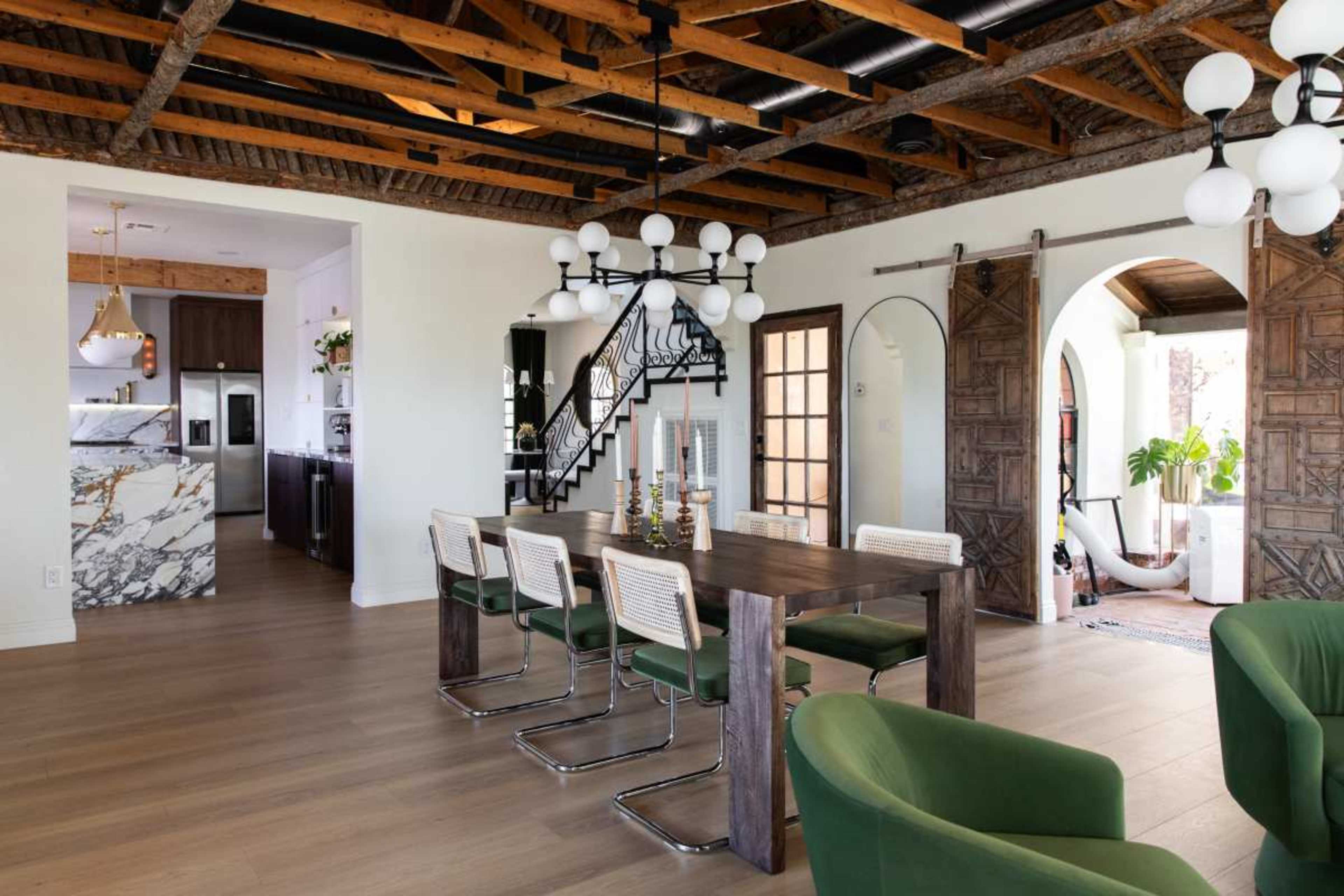 The image shows a modern dining area with a wooden table surrounded by green chairs, an open kitchen in the background, and decorative lighting overhead.
