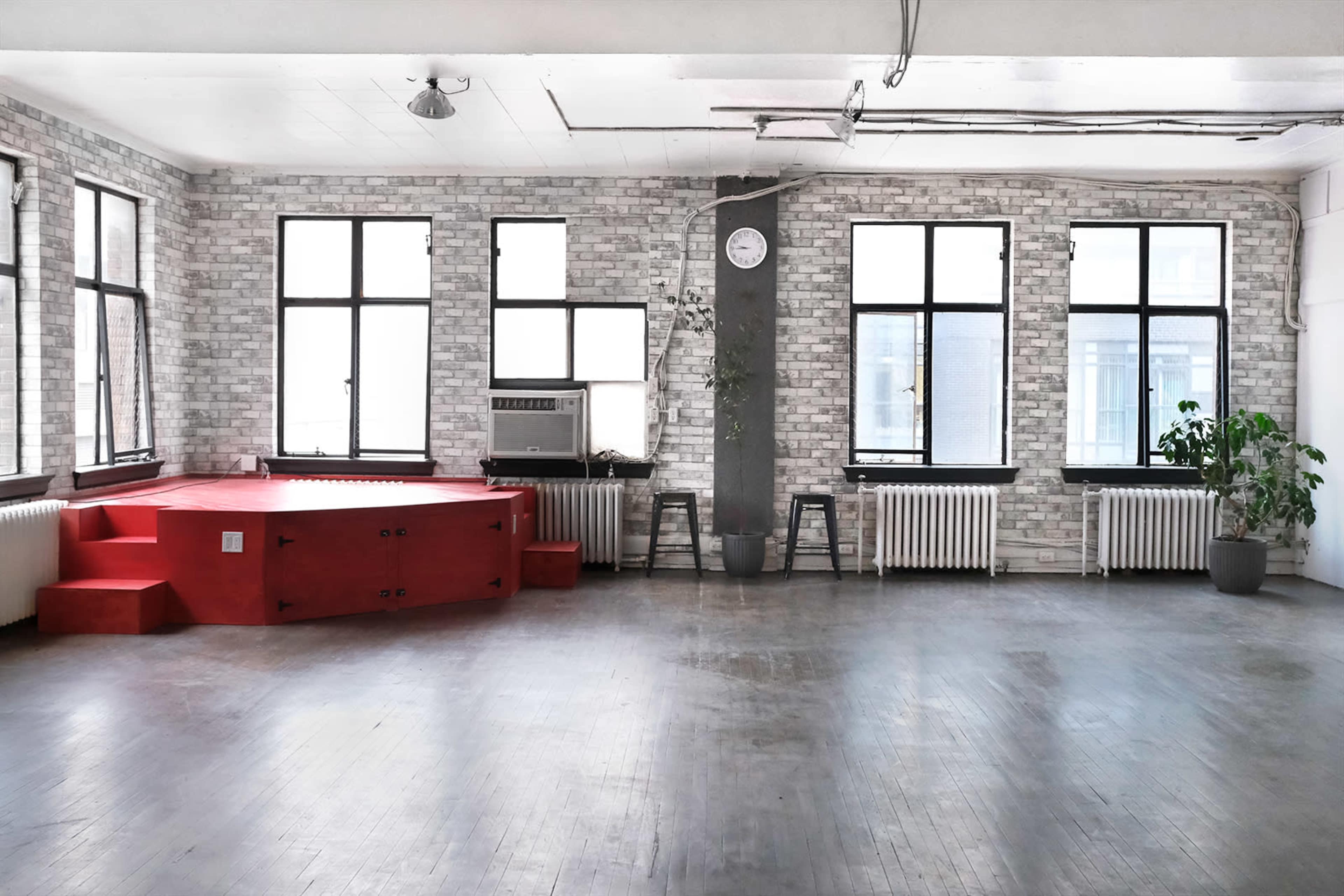 A spacious room with white brick walls, large windows, a red platform, and a couple of chairs against a backdrop of radiators.