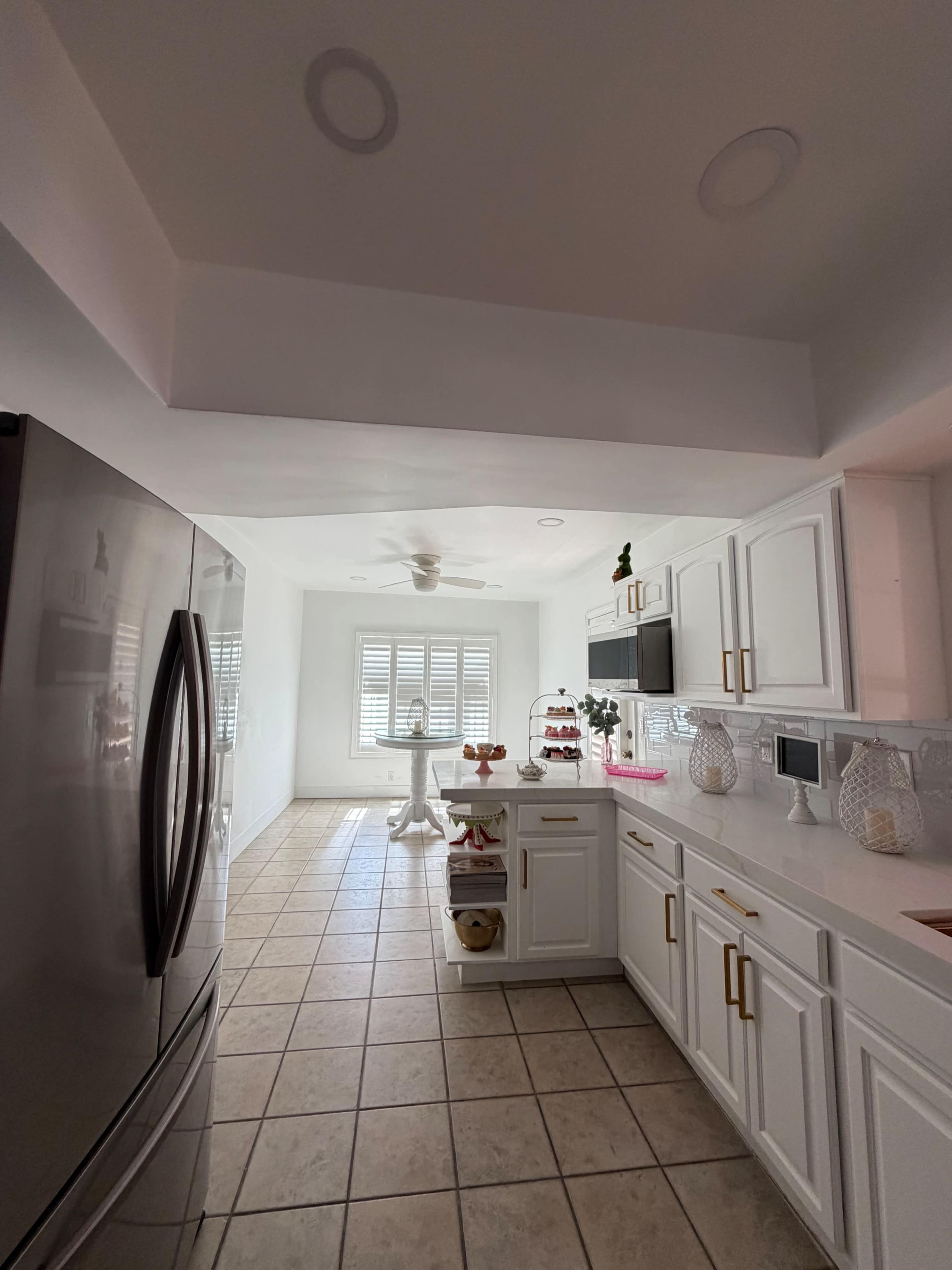 The kitchen features white cabinetry, a stainless steel refrigerator, and a tiled floor, leading to a brightly lit dining area with a ceiling fan and window shutters.