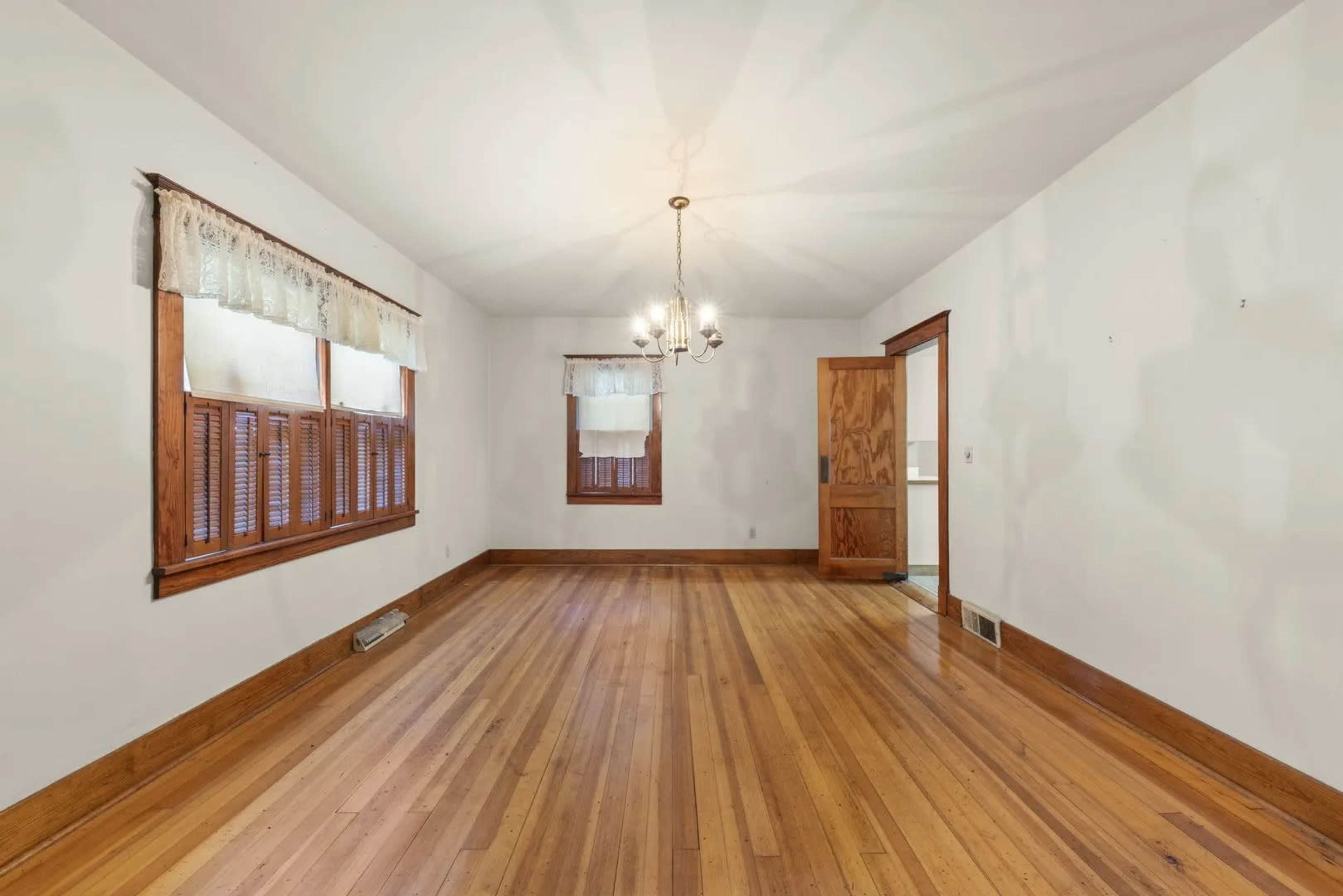 The image shows an empty room with wooden flooring, wooden window frames, and a chandelier hanging from the ceiling.