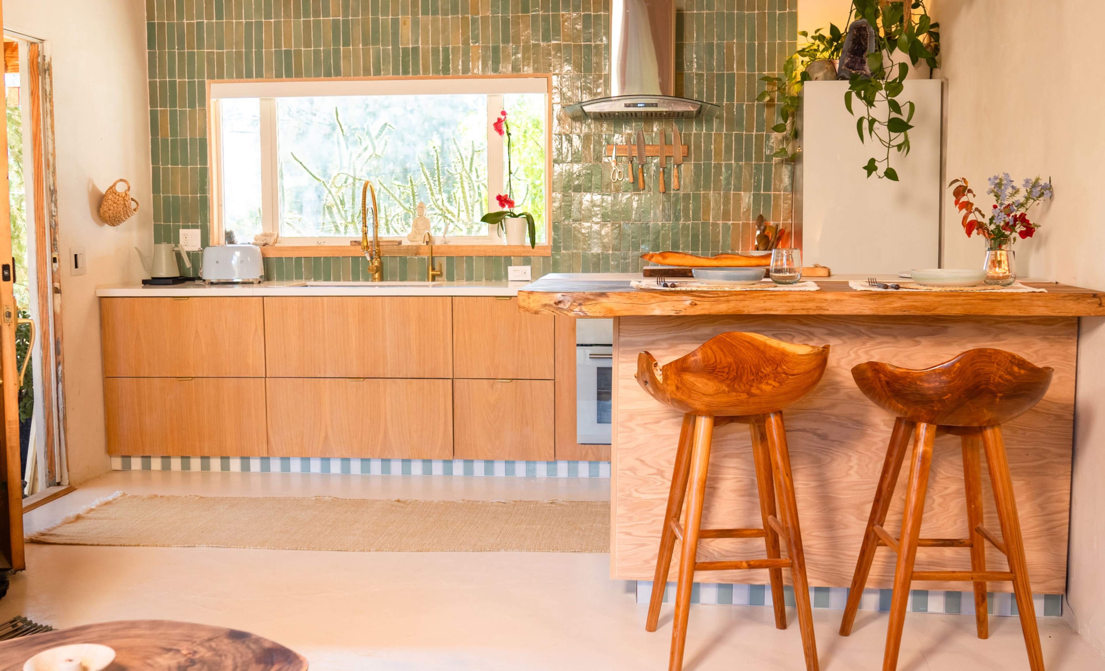 The image shows a modern kitchen with wooden cabinetry, a breakfast bar with two wooden stools, and green tiled walls.