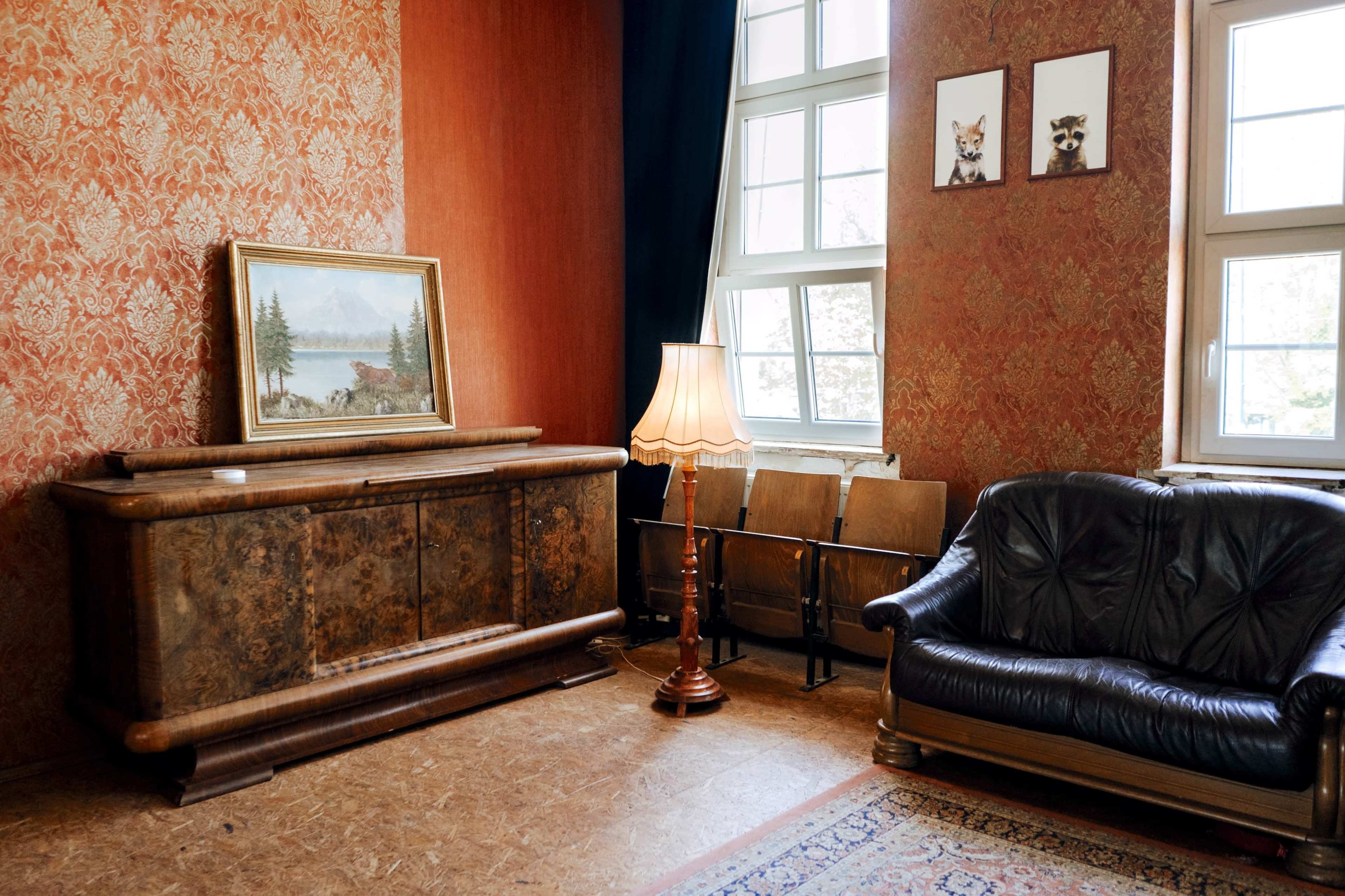 The room features a textured wallpaper, a wooden cabinet with a landscape painting above it, a standing lamp, and a black leather sofa.