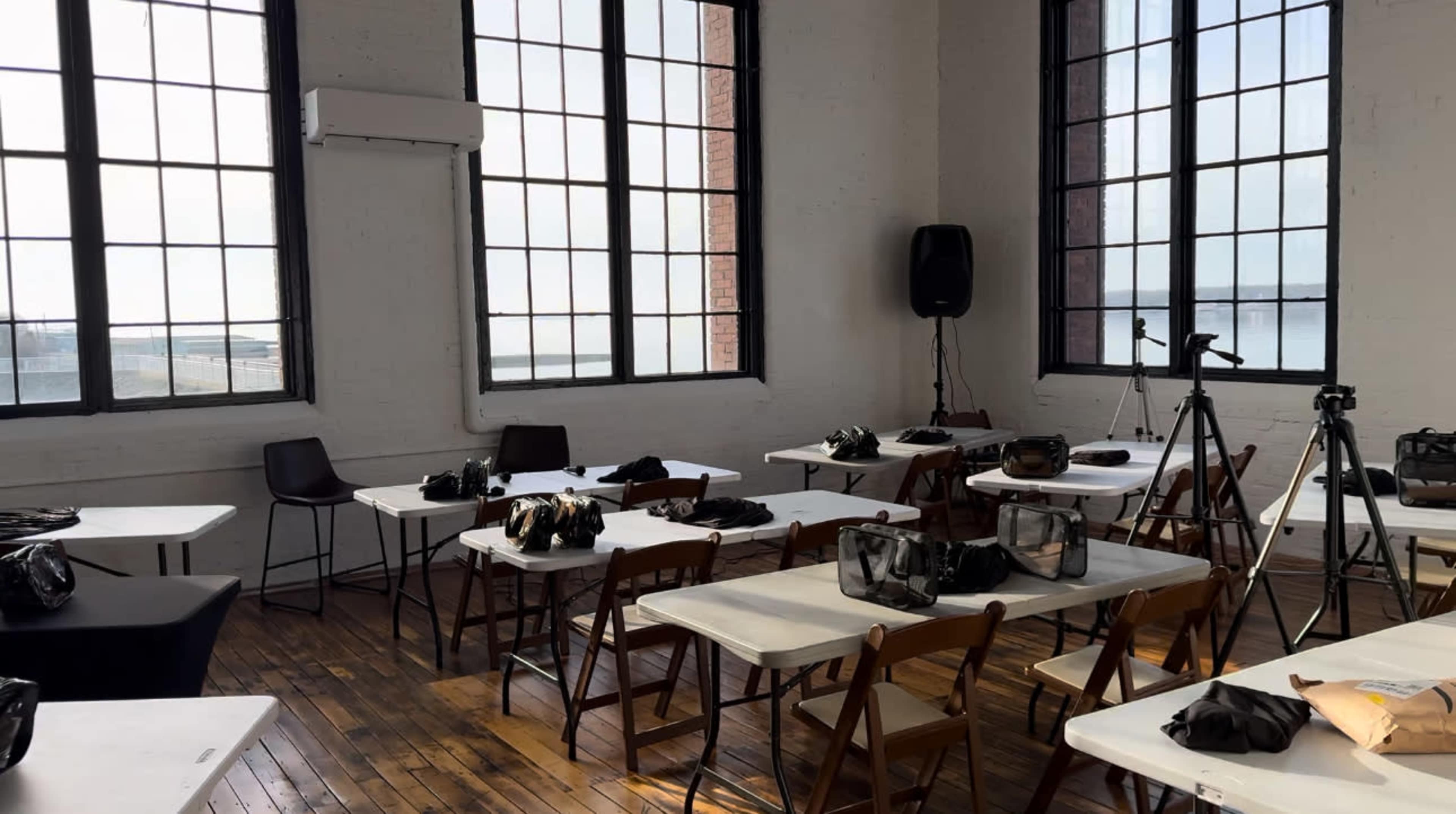 A room with large windows features several tables and chairs, each equipped with bags and cameras, indicating a setup for a workshop or event.