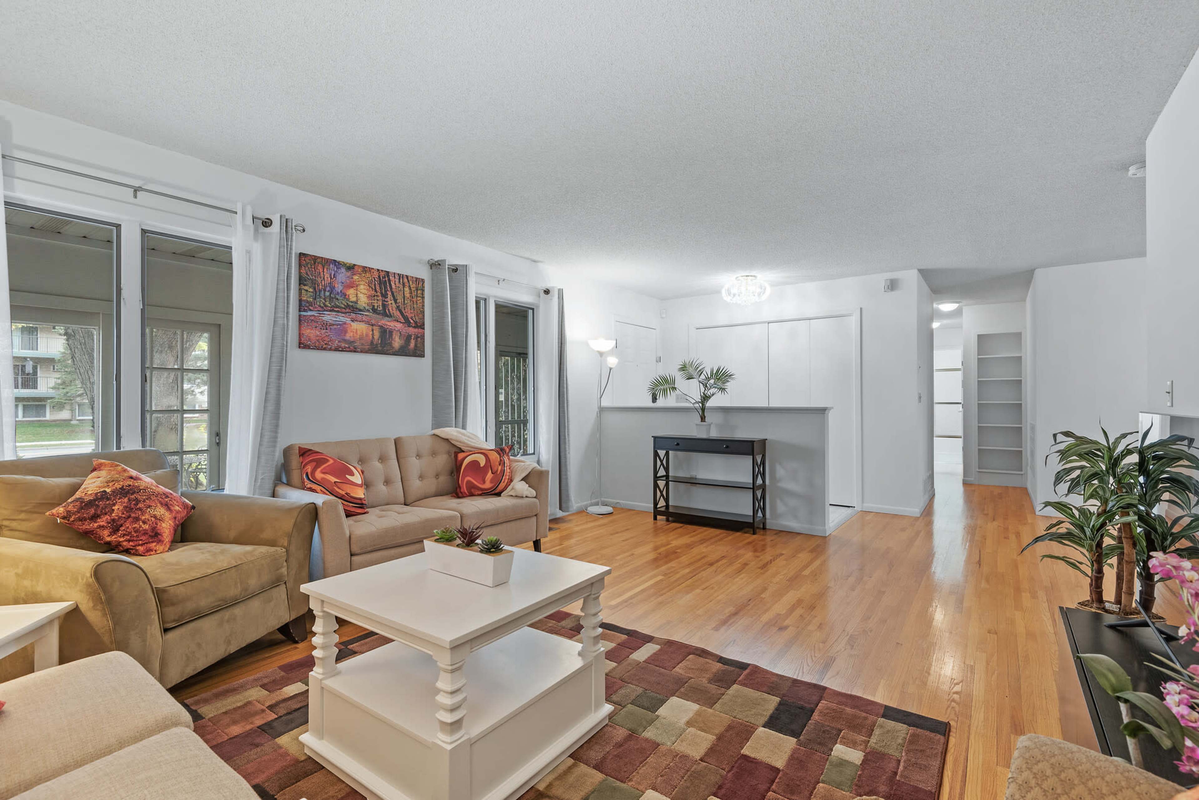The image shows a bright living room with two sofas, a coffee table, and a bar area, featuring wooden floors and large windows.
