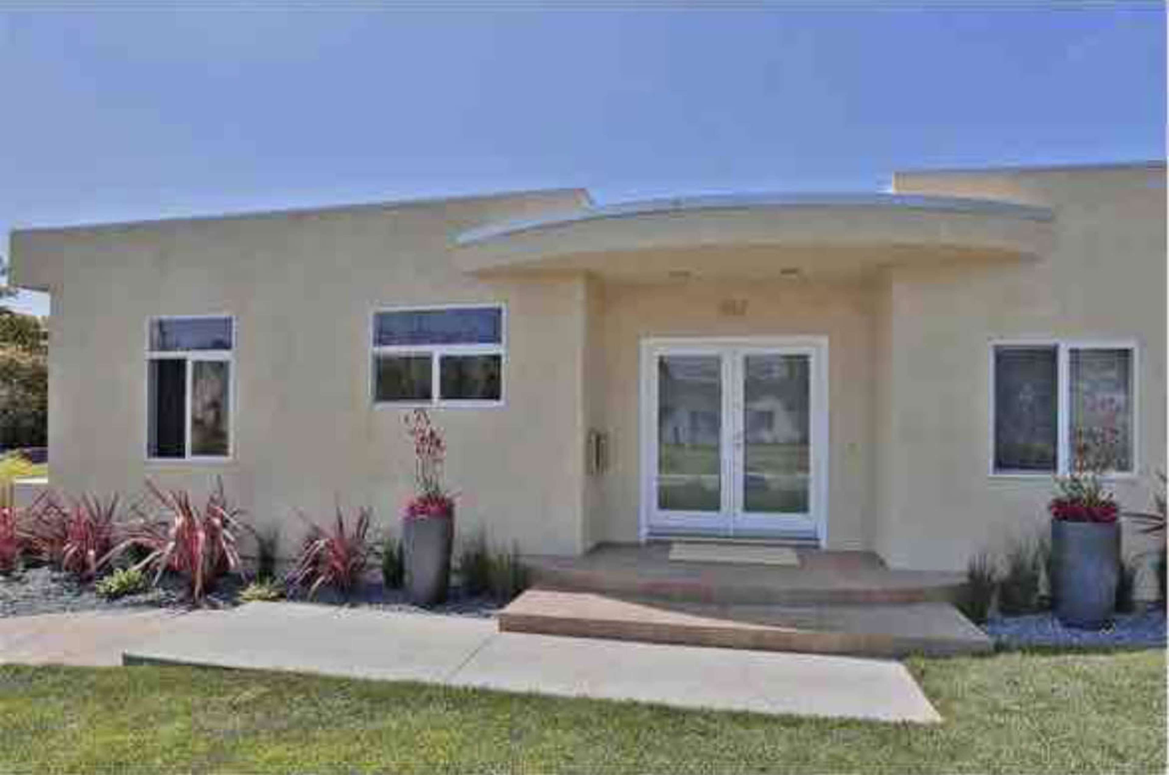 A modern, light-colored house with a curved entryway, large front windows, and potted plants flanking the entrance.