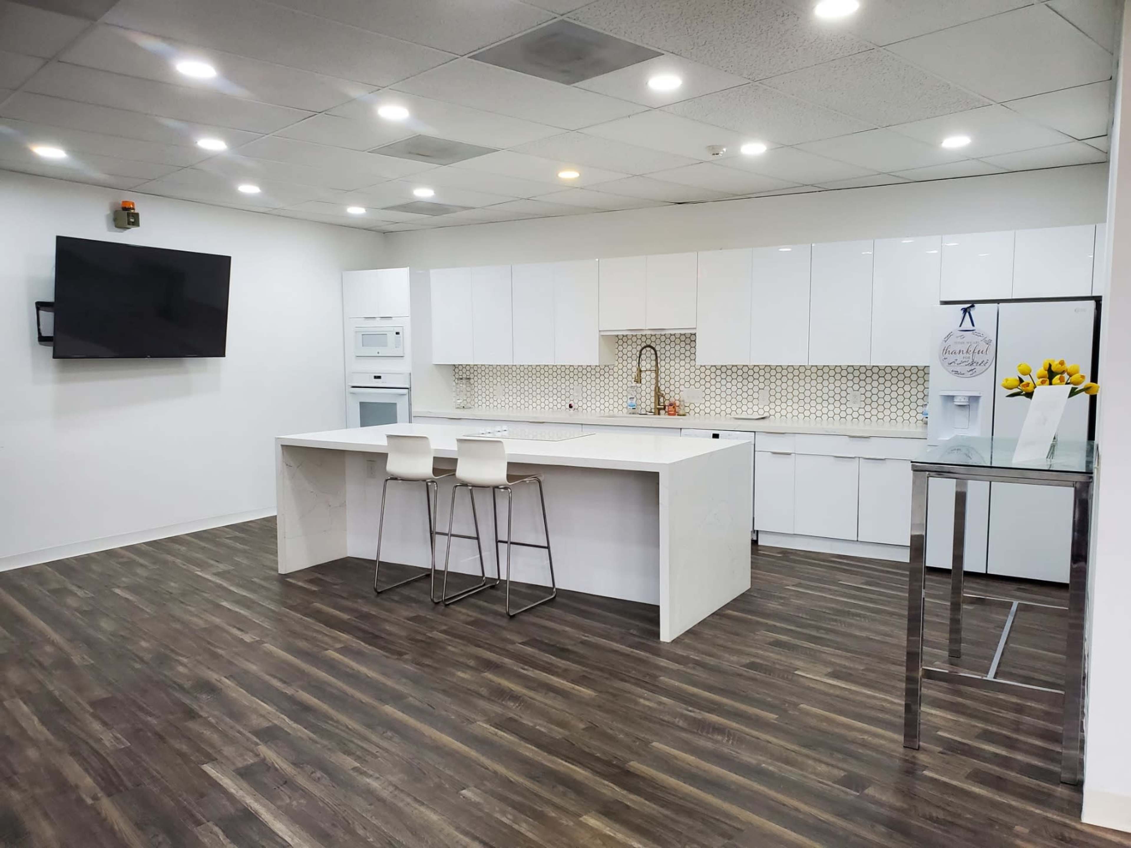 A modern kitchen with white cabinetry, a large island with two stools, and a wall-mounted television.