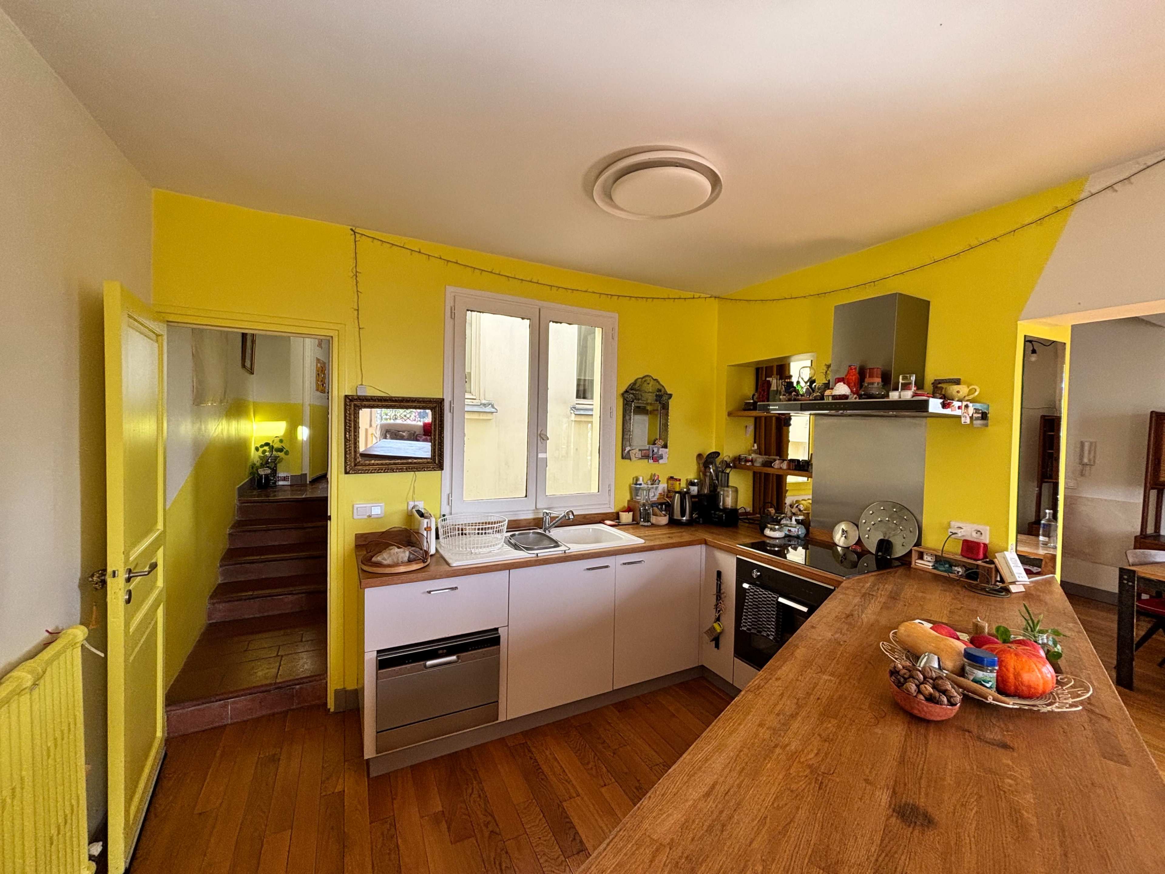 The image shows a bright yellow kitchen with modern appliances, a wooden counter, and a view of stairs leading to another room.