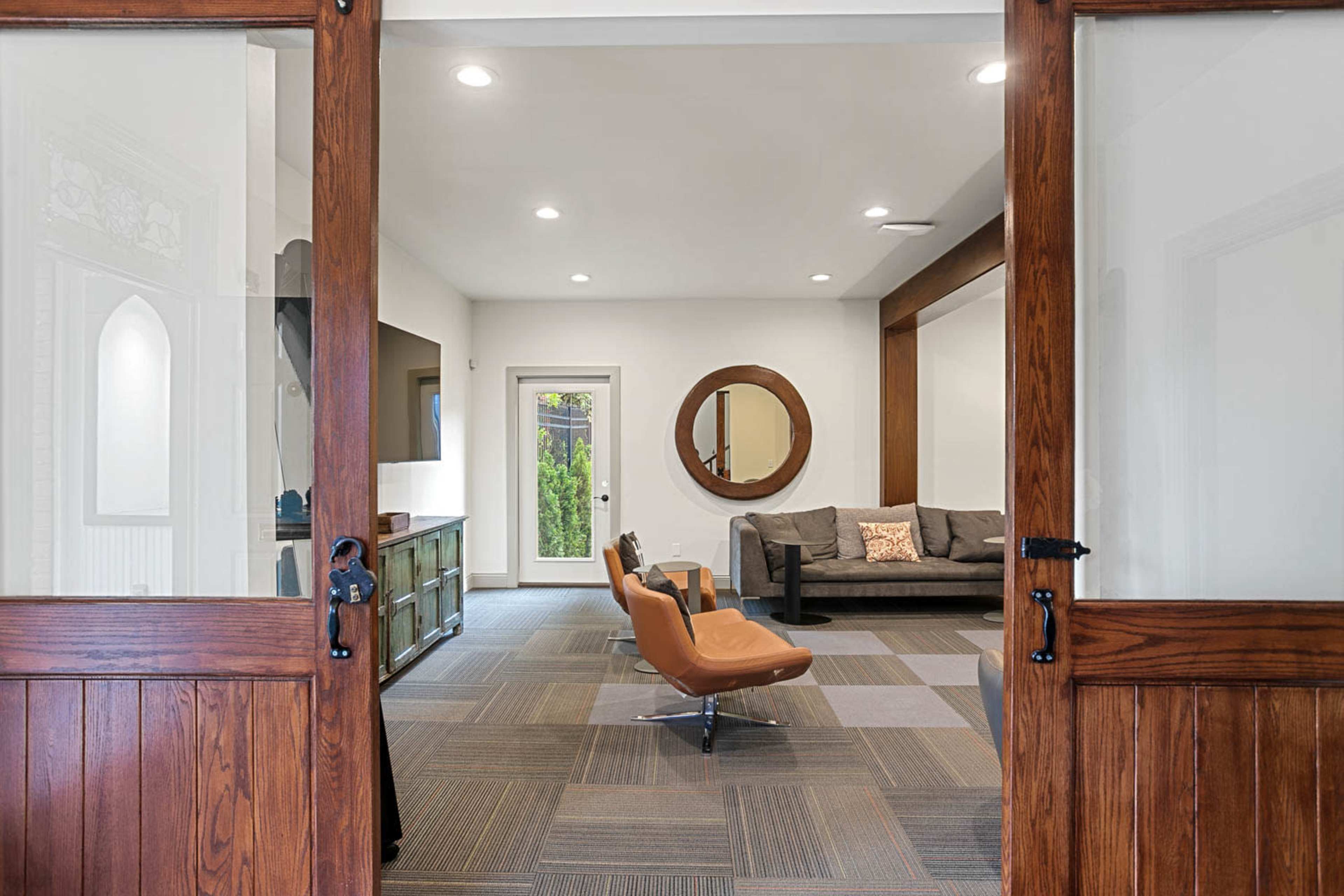 The image shows a seating area with a brown sofa, an orange chair, and a round mirror, visible through wooden double doors.